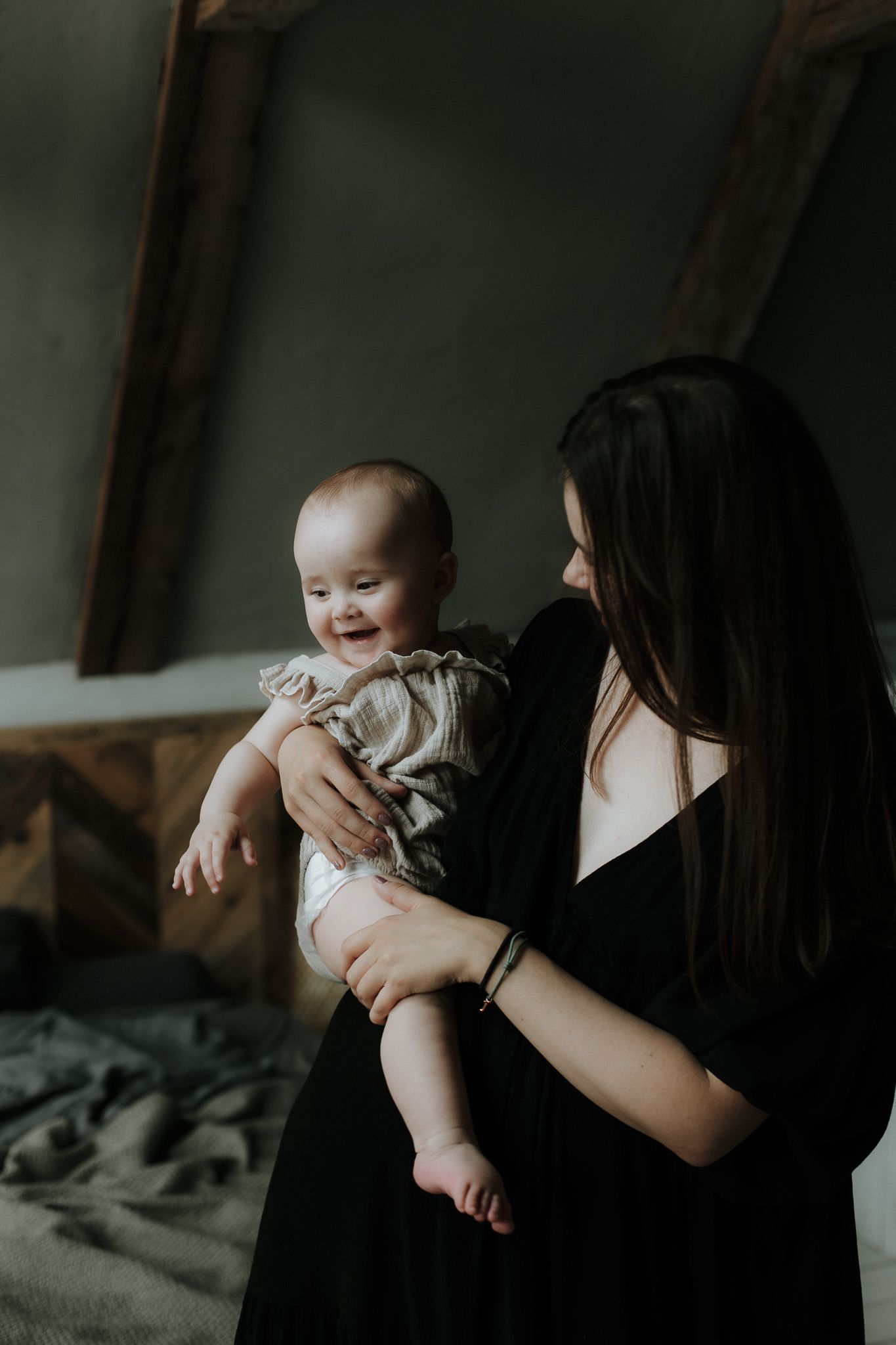 A dark-haired mother holds her smiling baby in a moody indoor setting.