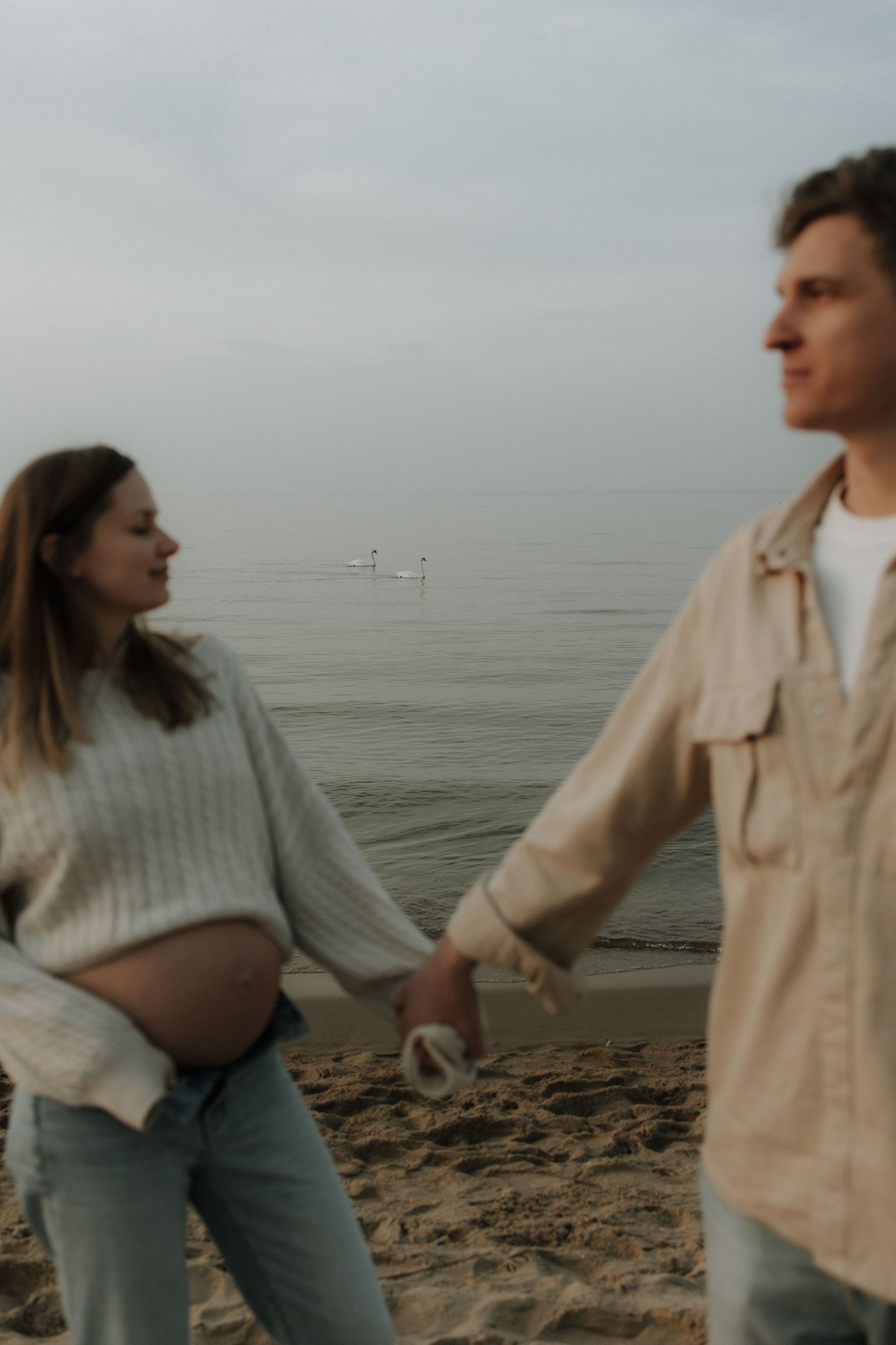 Pregnant couple holding hands and walking on a sandy beach near calm ocean waters on an overcast day.
