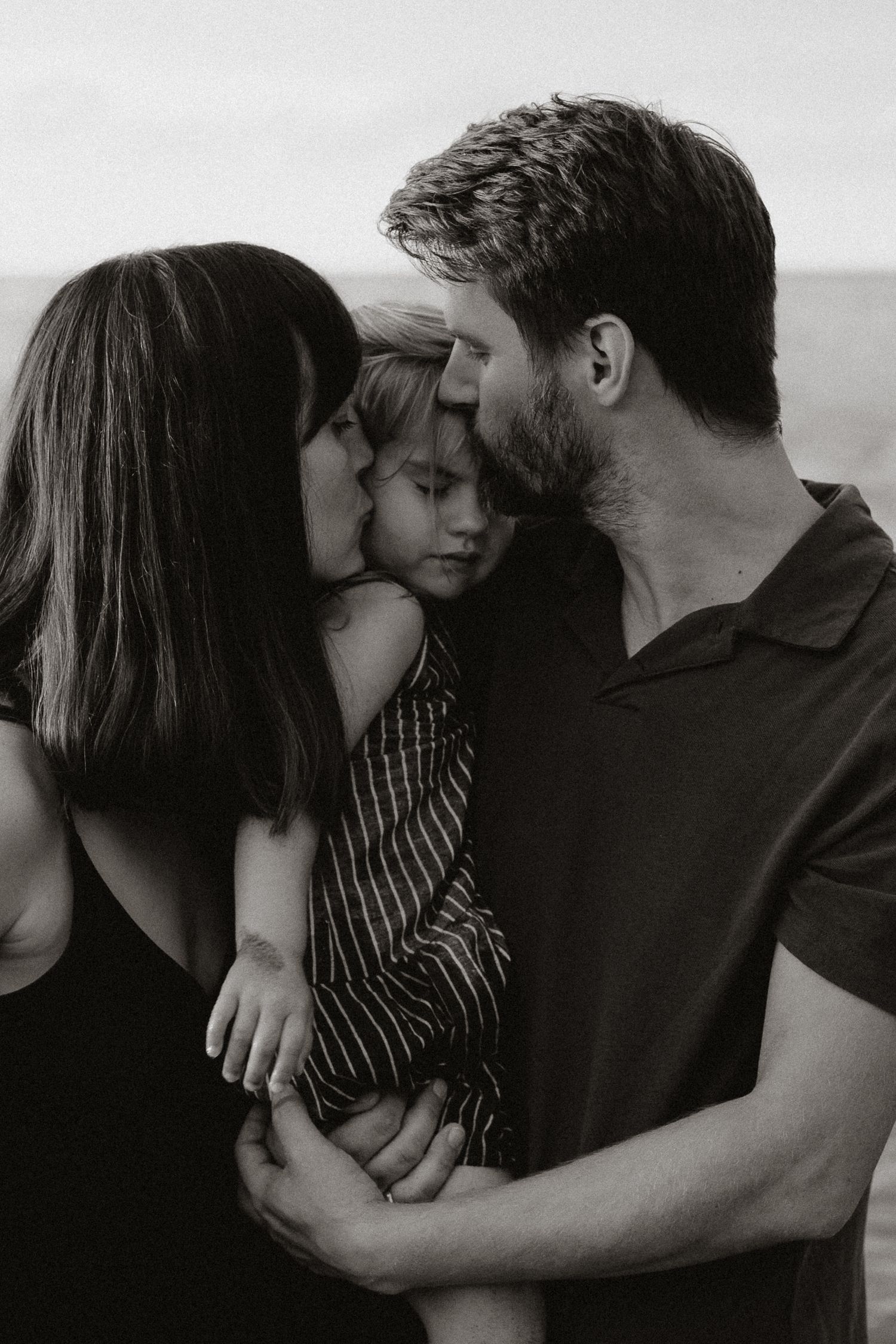 Black and white photo of parents kissing their young child on a beach.