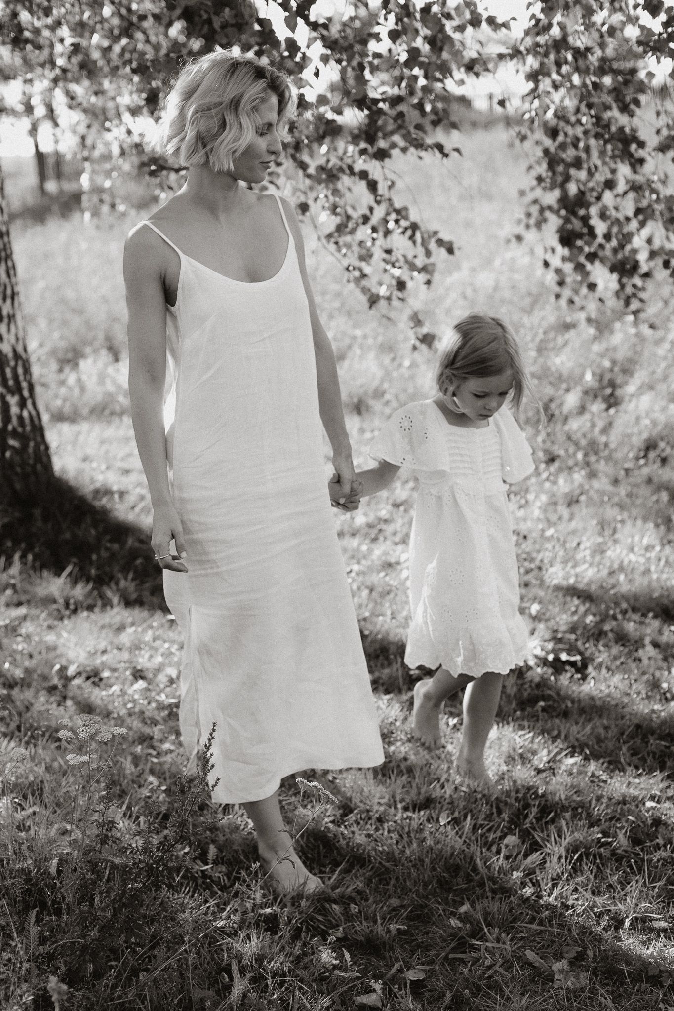 A mother and daughter in white dresses walk barefoot under a tree in black and white.