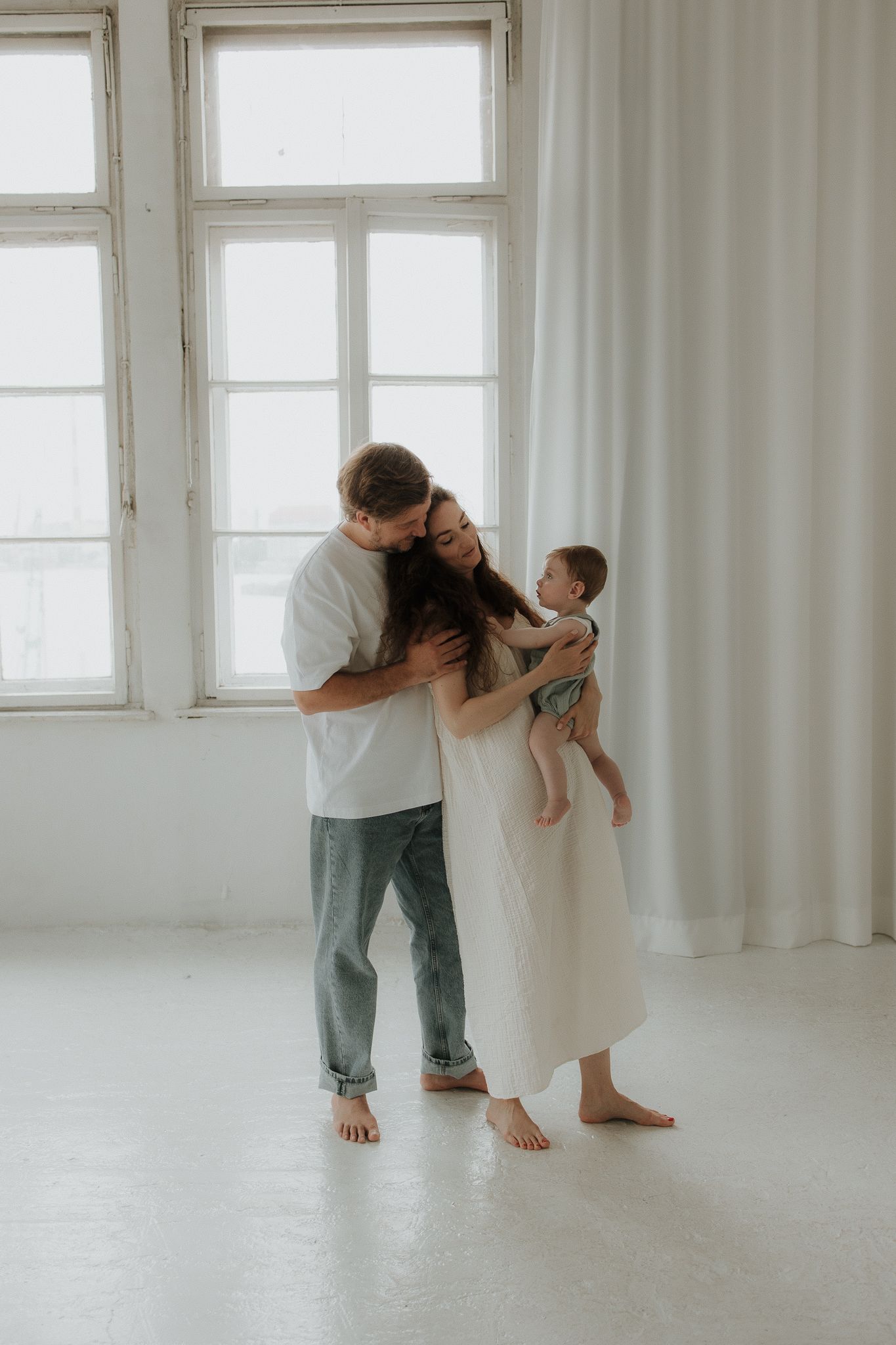 Young family with baby sharing a tender embrace in a bright white minimalist room.