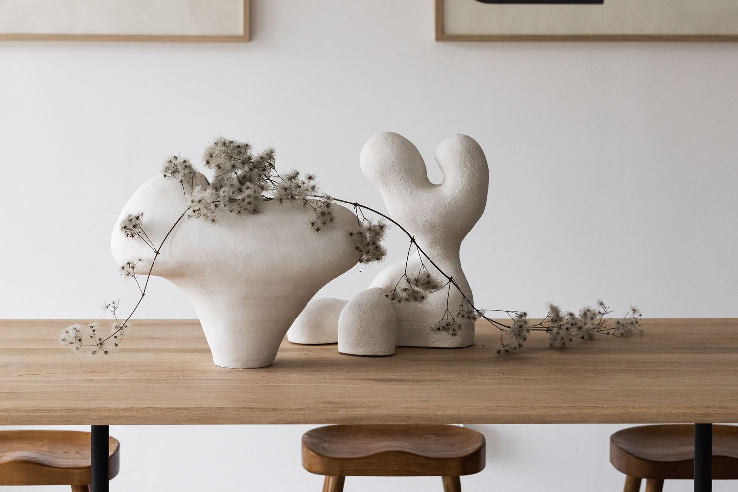 White ceramic sculptural vases with dried flowers on a wooden dining table, minimalist Scandinavian interior decor.