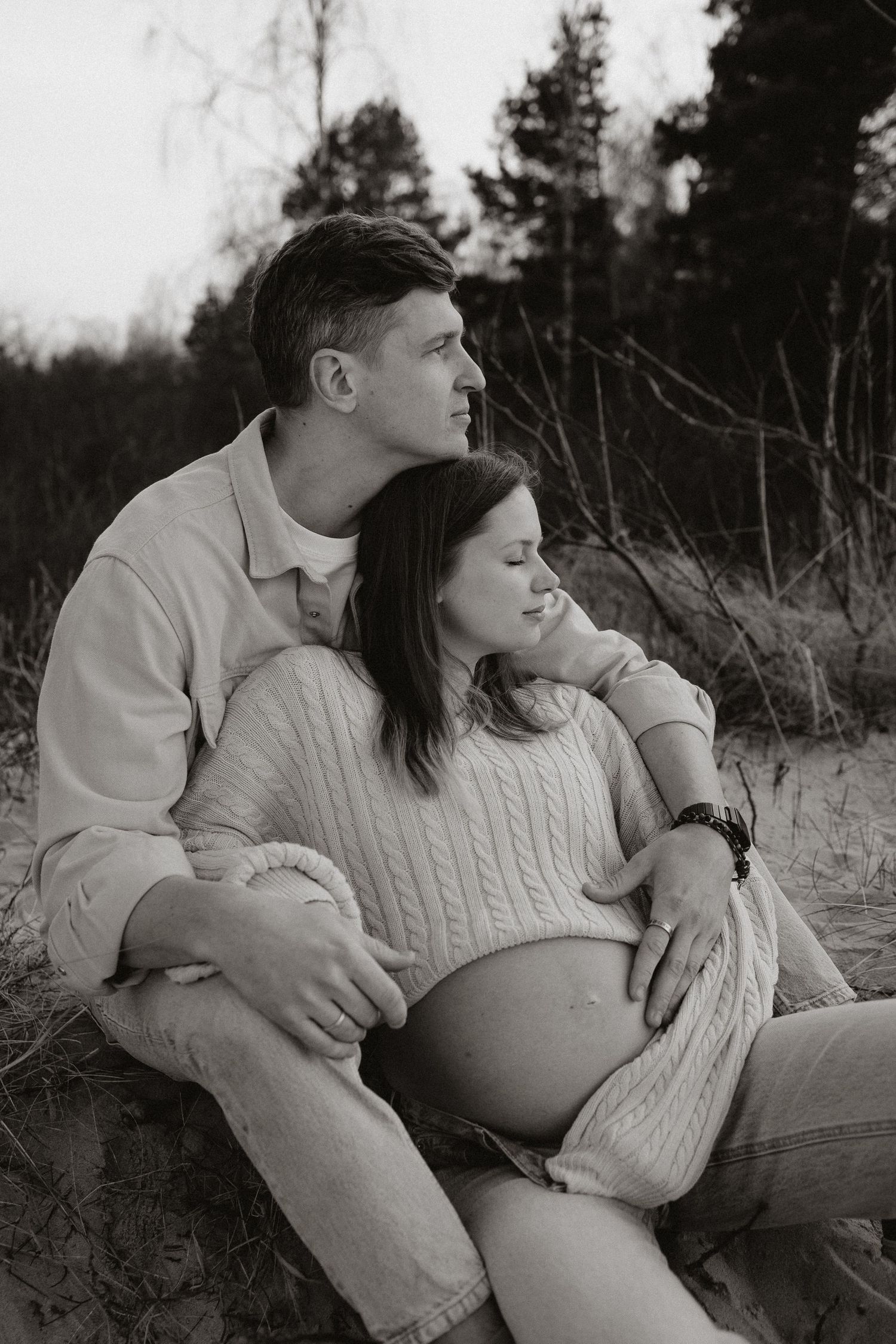 Black and white photo of couple sitting in sand dunes, man embracing pregnant partner.