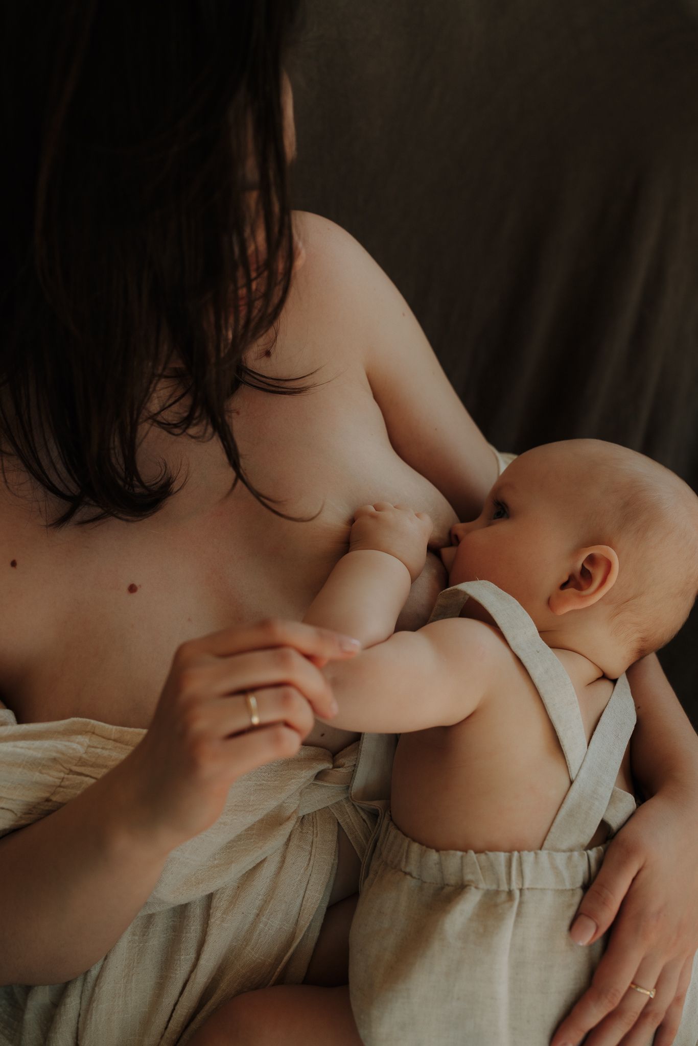 Mother breastfeeding her infant baby dressed in linen overalls in a warm, intimate setting.