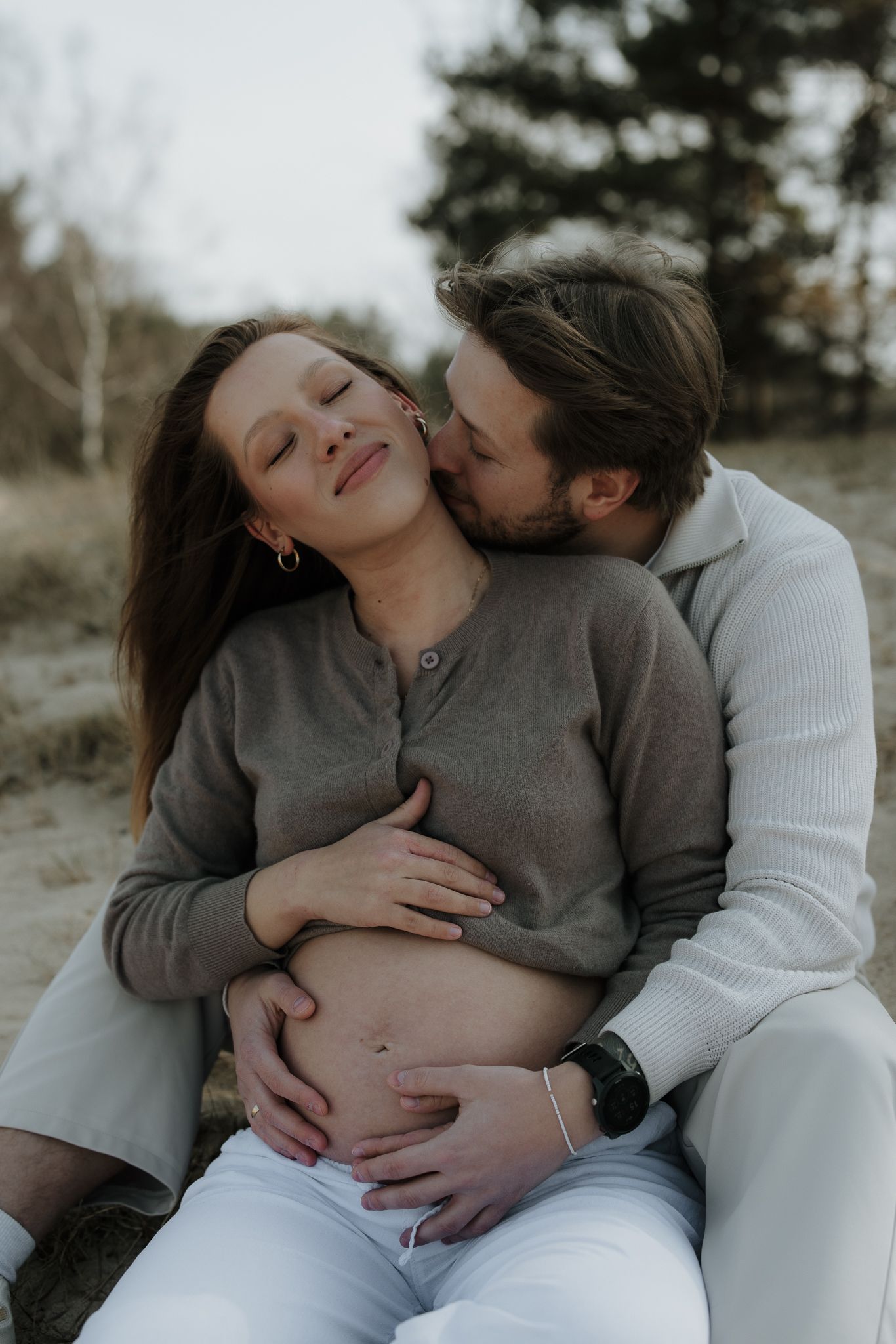 Man kissing pregnant woman's cheek while embracing her baby bump on sandy beach.