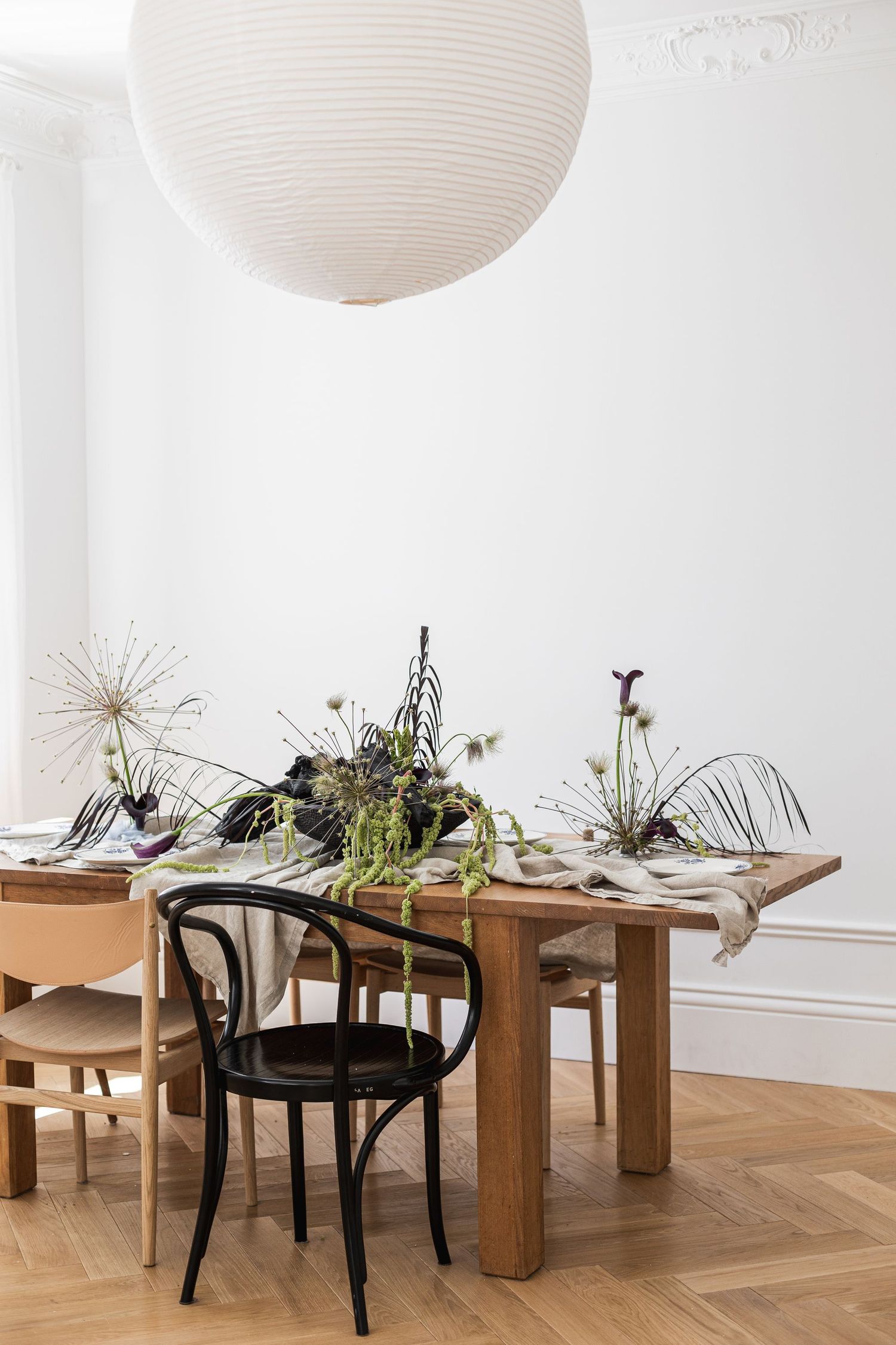 Minimalist dining room with wooden table, black Thonet chair, botanical centerpiece, and white paper lantern pendant light.