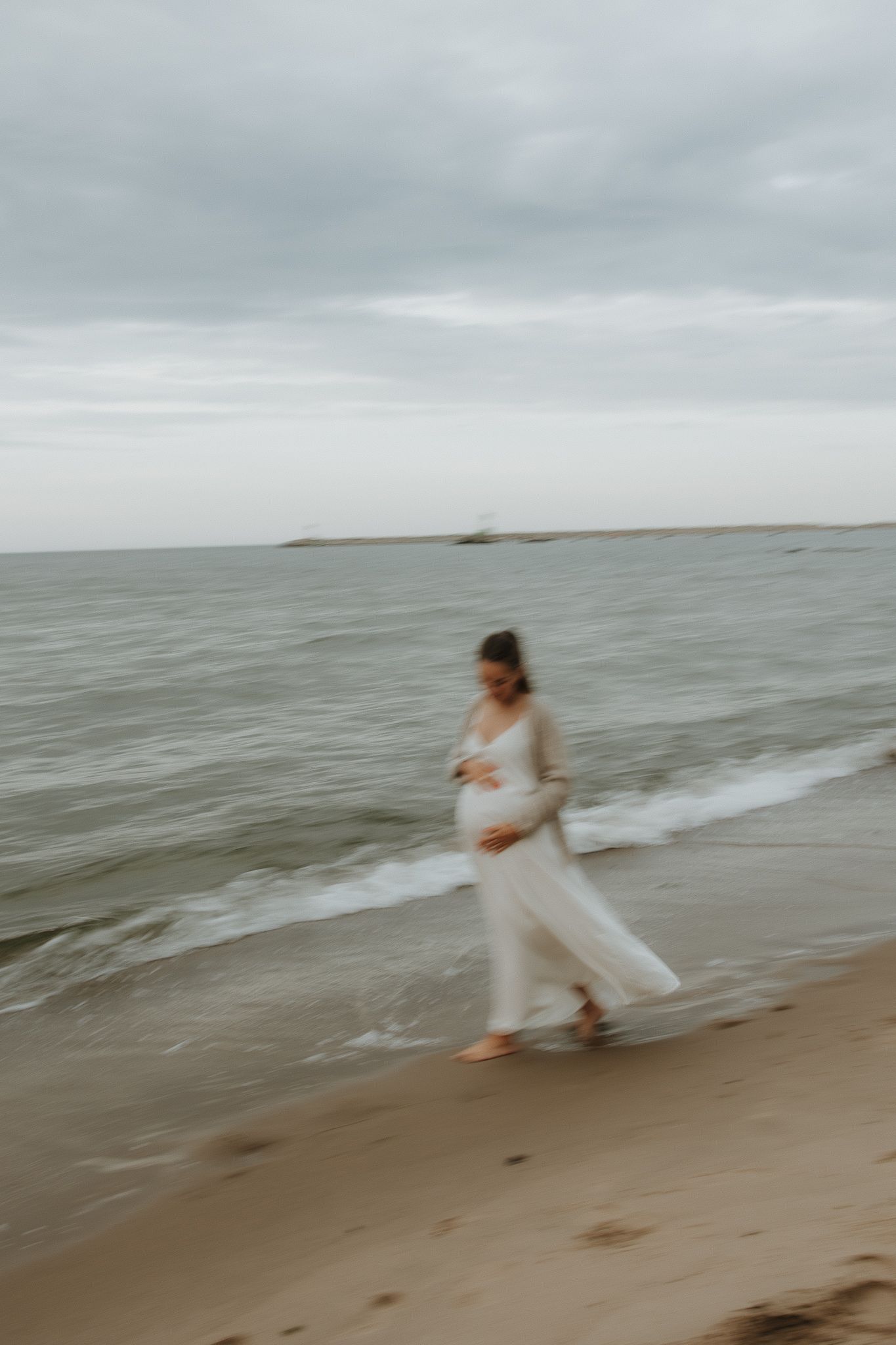 Pregnant woman in white dress walks along a sandy beach with ocean waves behind her.