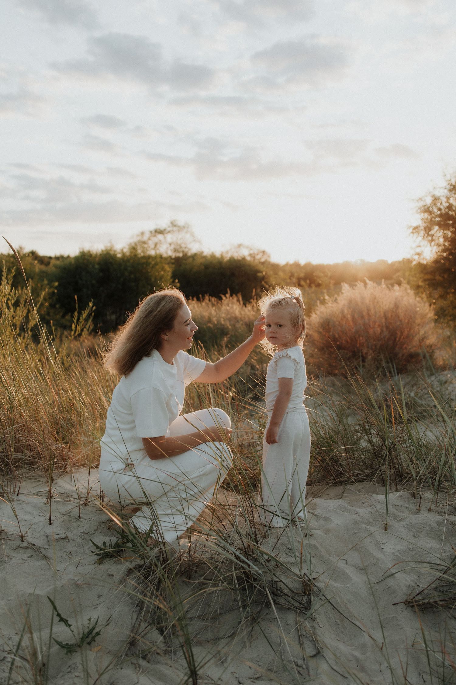 Mother in white outfit kneeling on sand dunes gently touching her toddlers face at golden sunset.