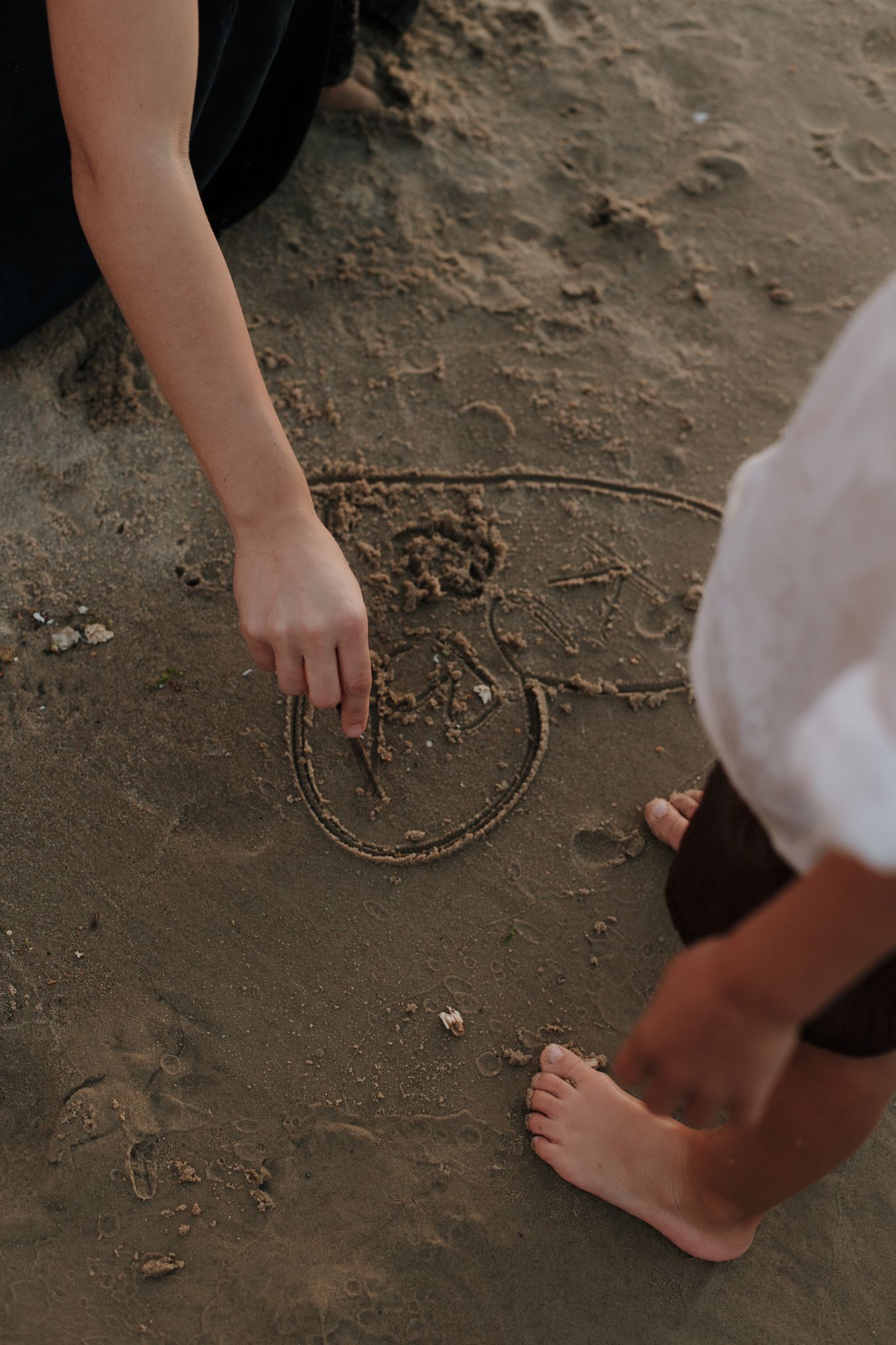 A person draws a smiley face in wet sand on a beach with bare feet nearby.