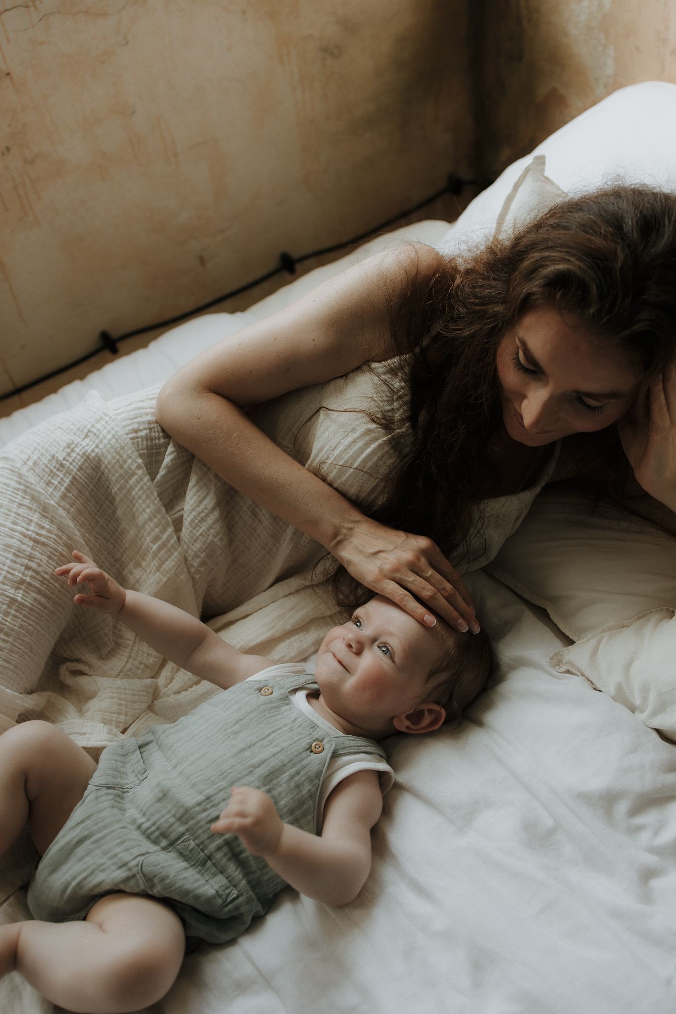 Mother lying on bed gently touching her smiling baby dressed in sage green romper.