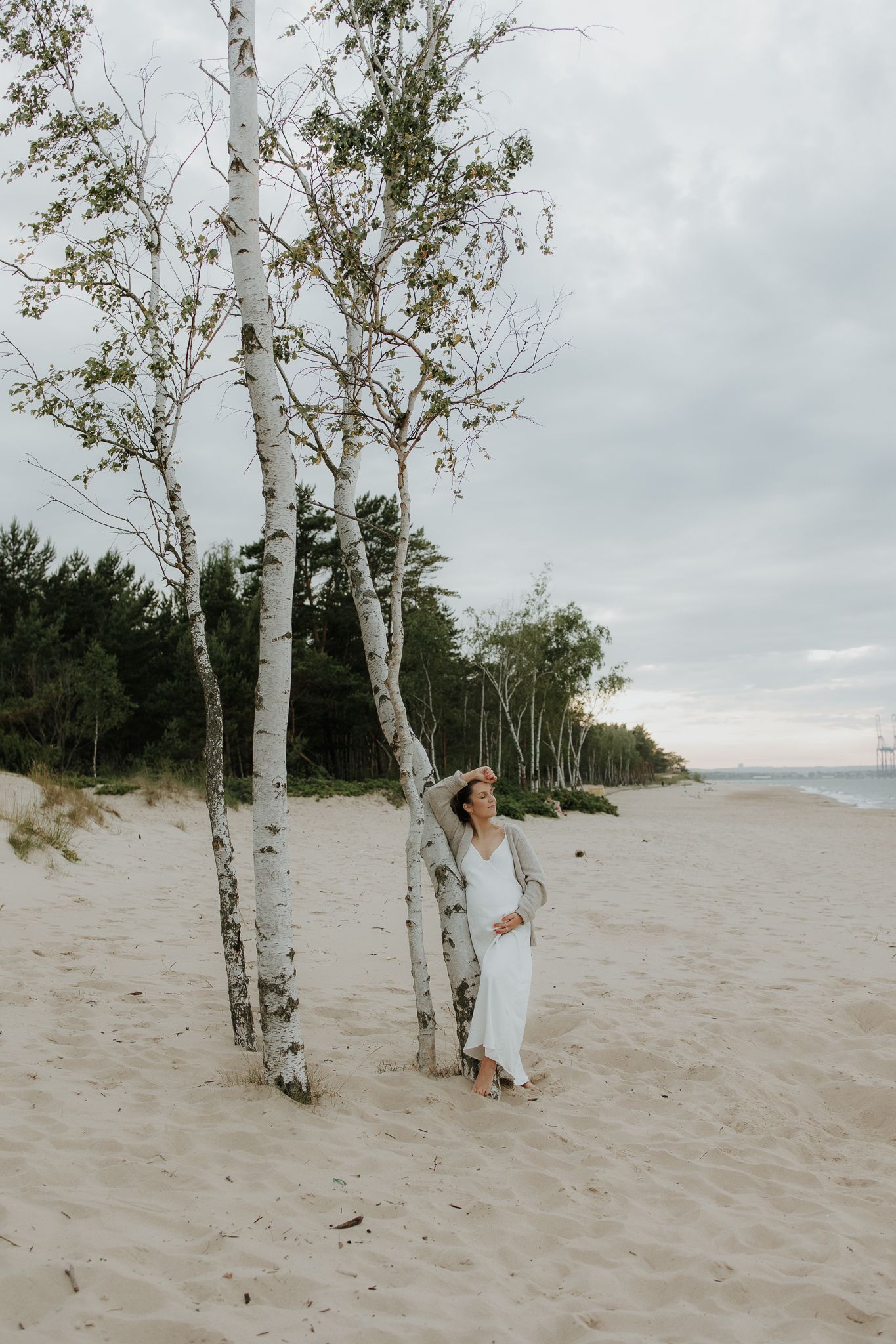 Woman in white dress posing by birch trees on a sandy beach with overcast skies.