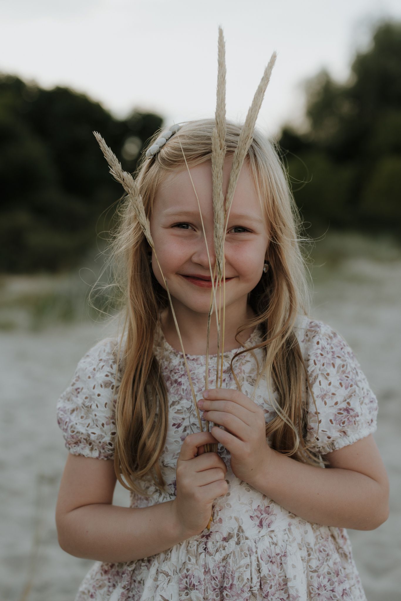 Smiling blonde girl in floral dress holding dried grass stems in front of her face.