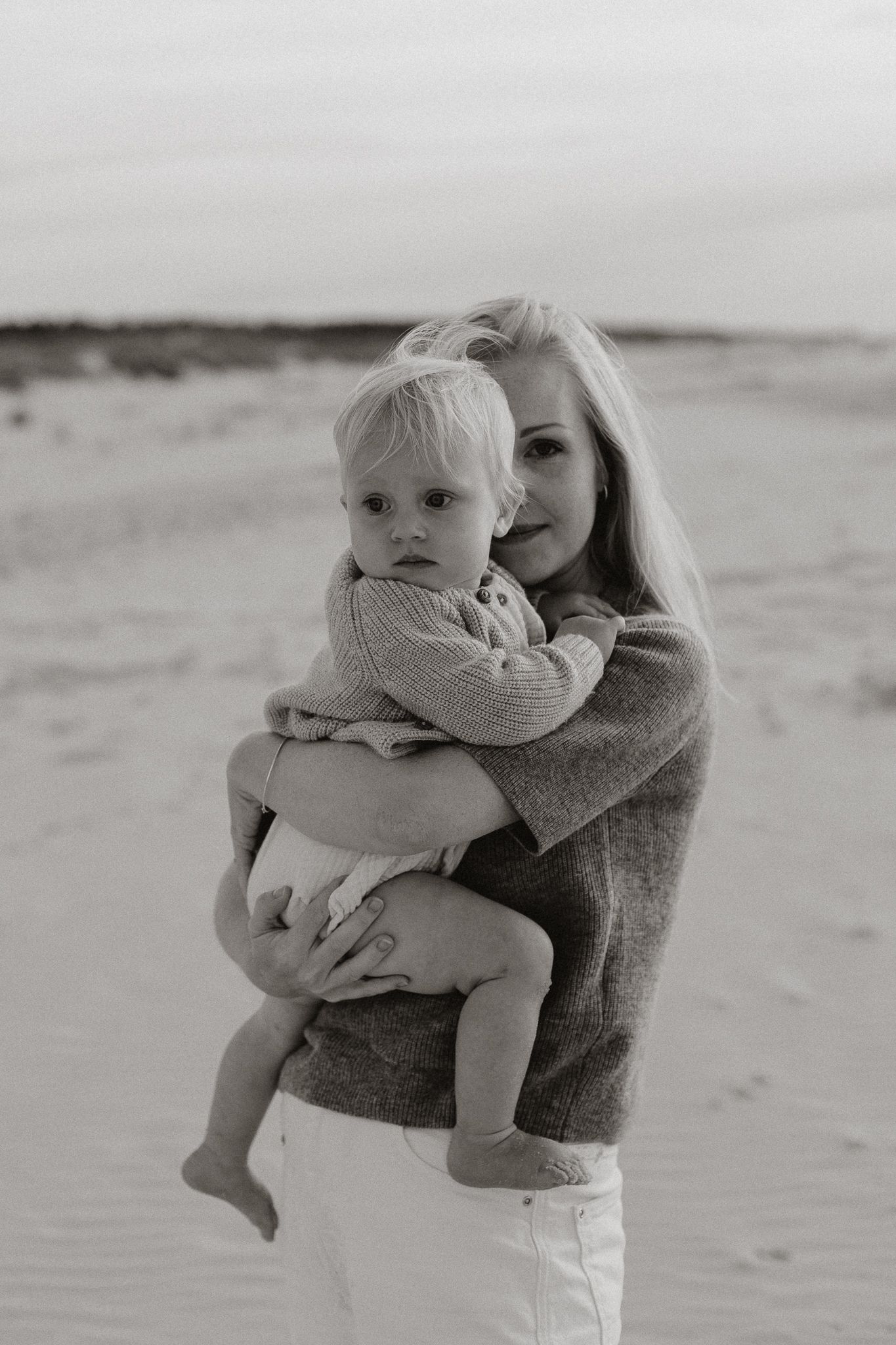 Black and white photo of blonde mother holding toddler on sandy beach at dusk.