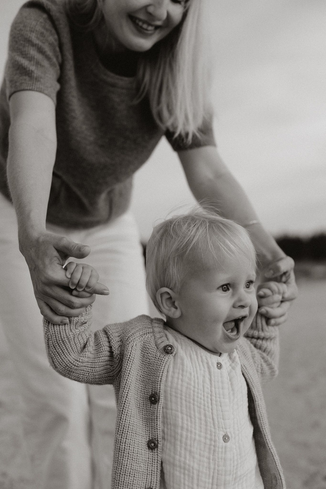Black and white photo of a laughing toddler being helped to walk by a smiling blonde mother.