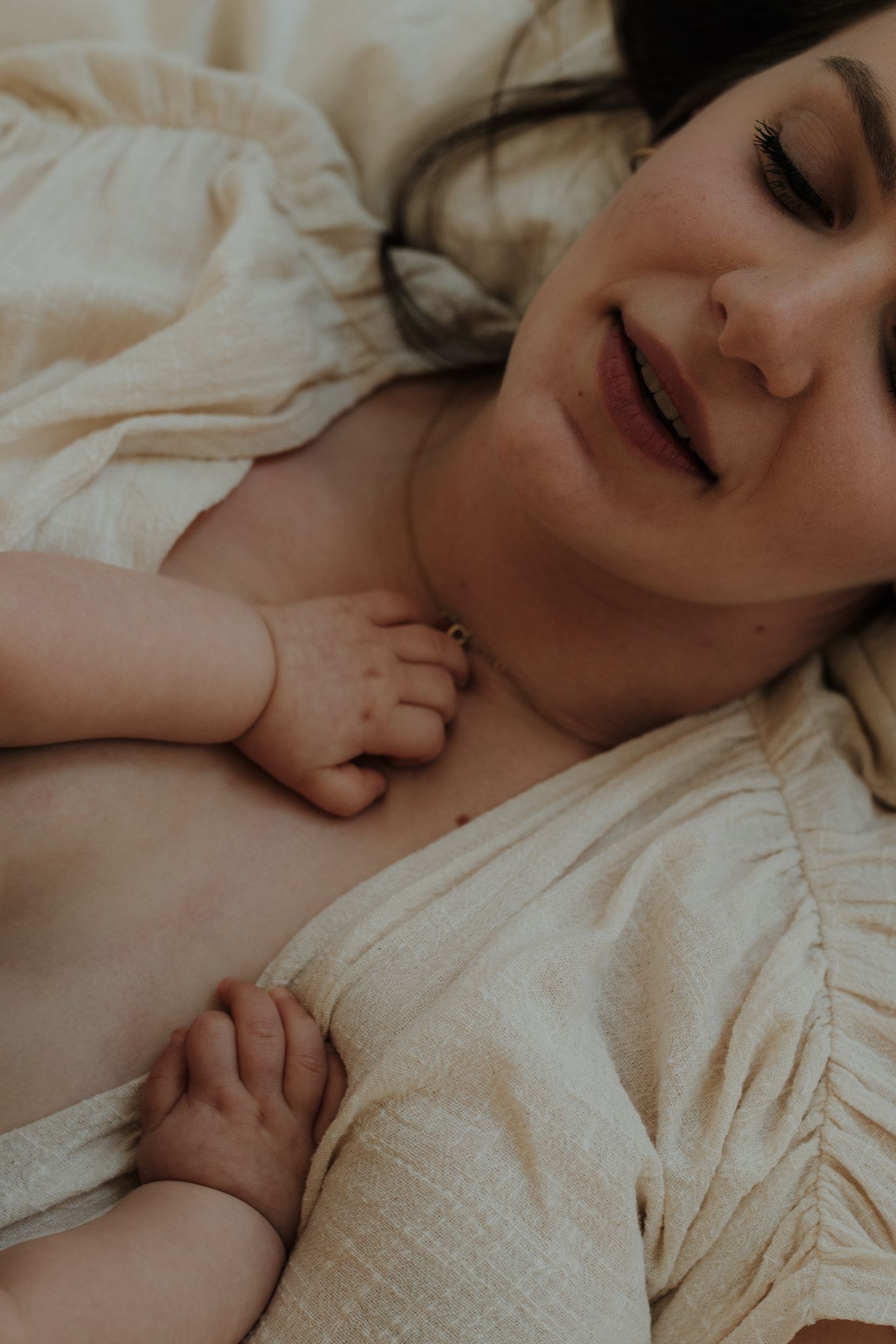 Smiling mother lying down with her newborn baby resting on her chest in soft linen bedding.