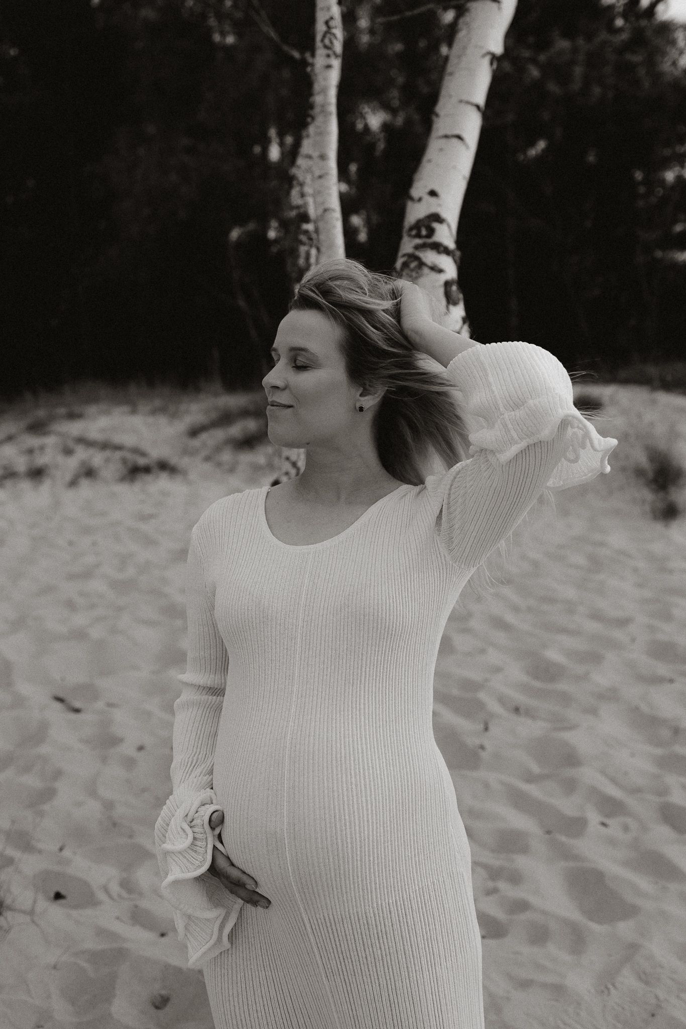 Black and white photo of pregnant woman in white ribbed dress posing near birch trees on sand.