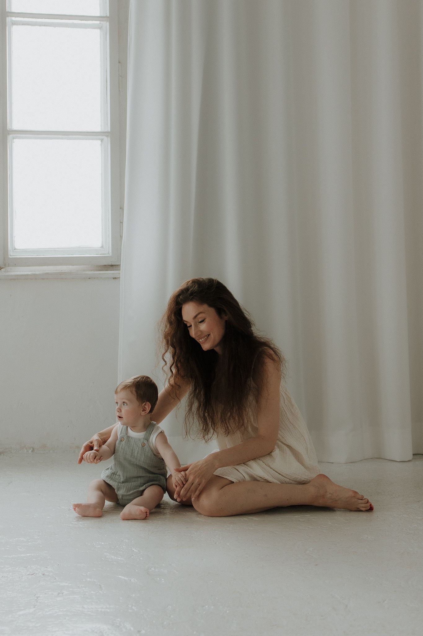 Young mother in white dress sitting on floor with baby in sage green romper near bright window.