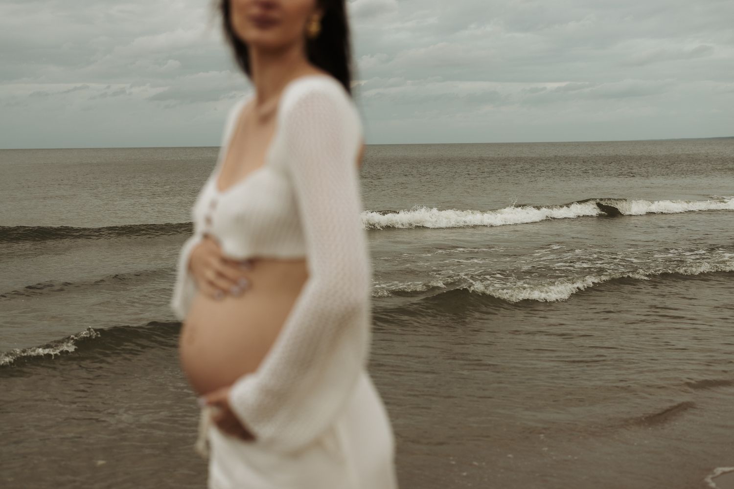 Blurred pregnant woman in white dress holding her baby bump on a cloudy beach.