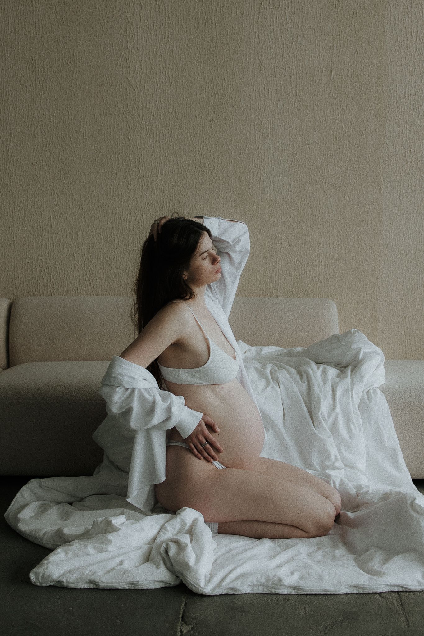 Pregnant woman in white lingerie and open shirt kneeling beside a beige sofa with white sheets.