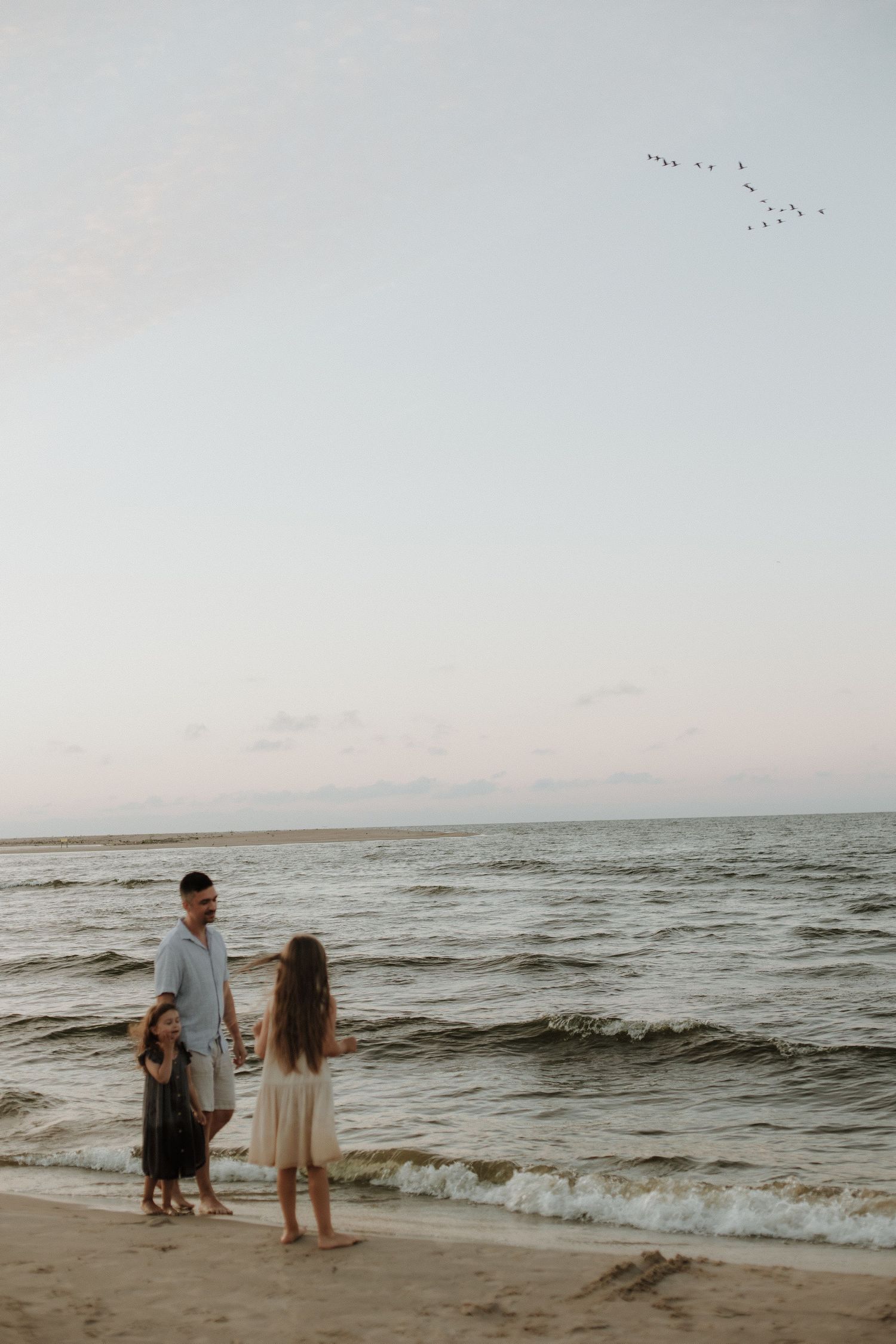 Father stands with two young daughters at sunset on a calm ocean beach.