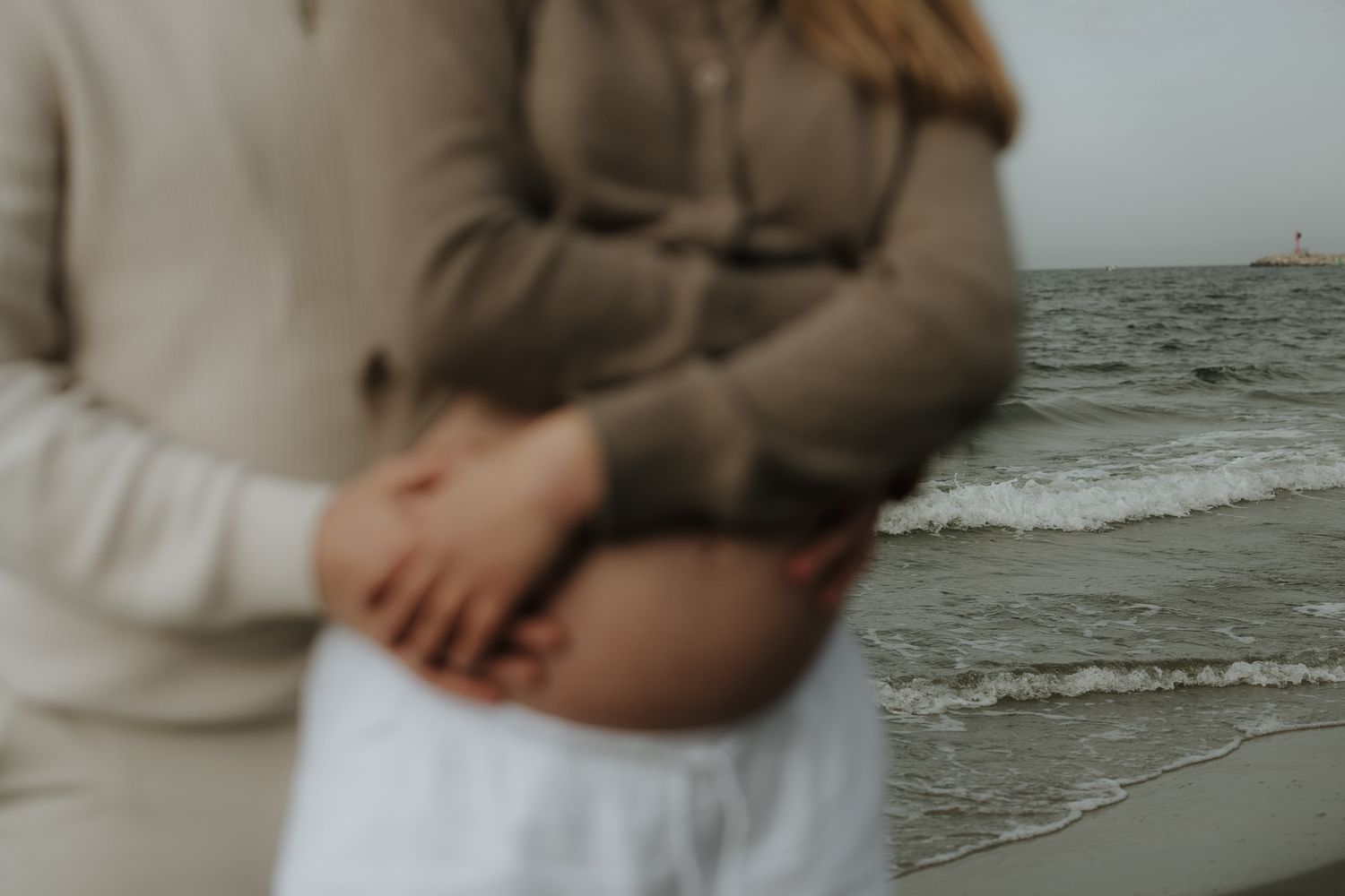 Close-up of couple holding pregnant belly near ocean waves, soft focus maternity photo.
