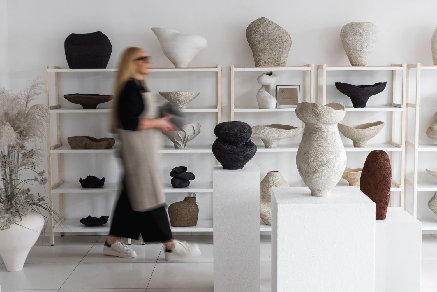 Woman walking past white shelves displaying ceramic sculptures and vases in a minimalist art gallery.