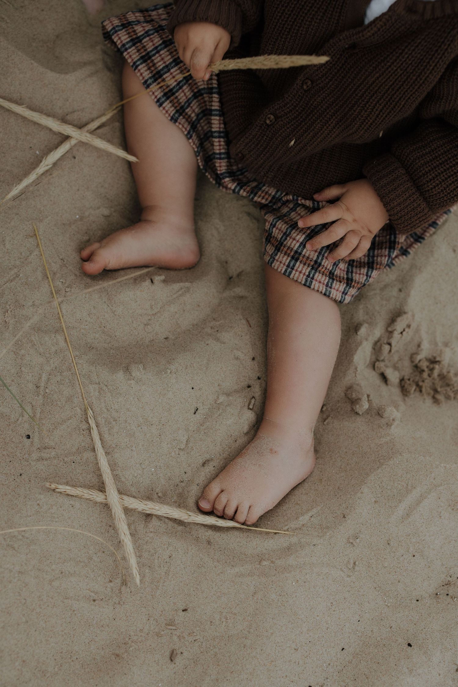 Close-up of toddler bare feet sitting on sand in plaid skirt and brown sweater.