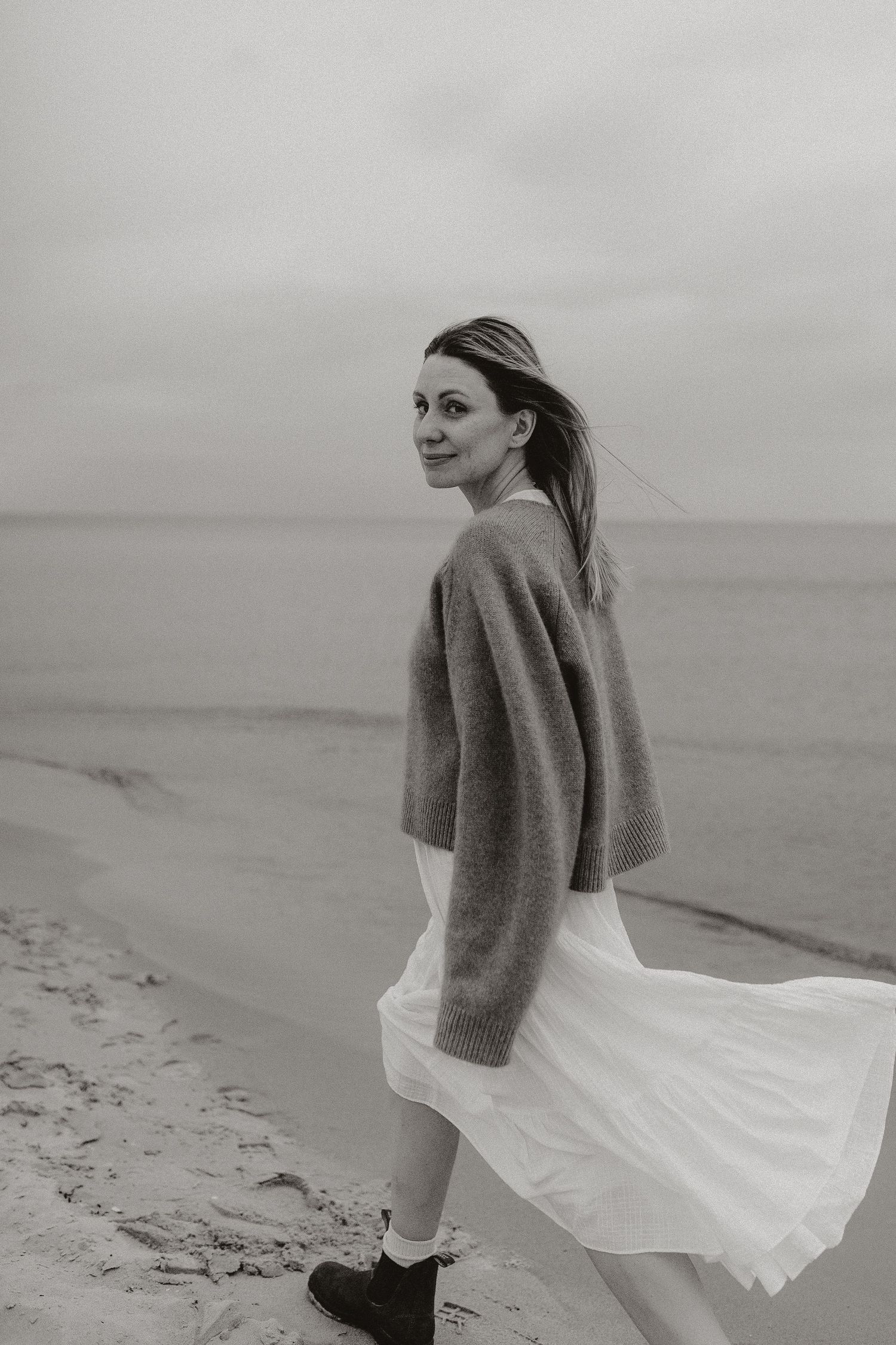 Black and white portrait of woman in white skirt and sweater walking on beach shoreline.