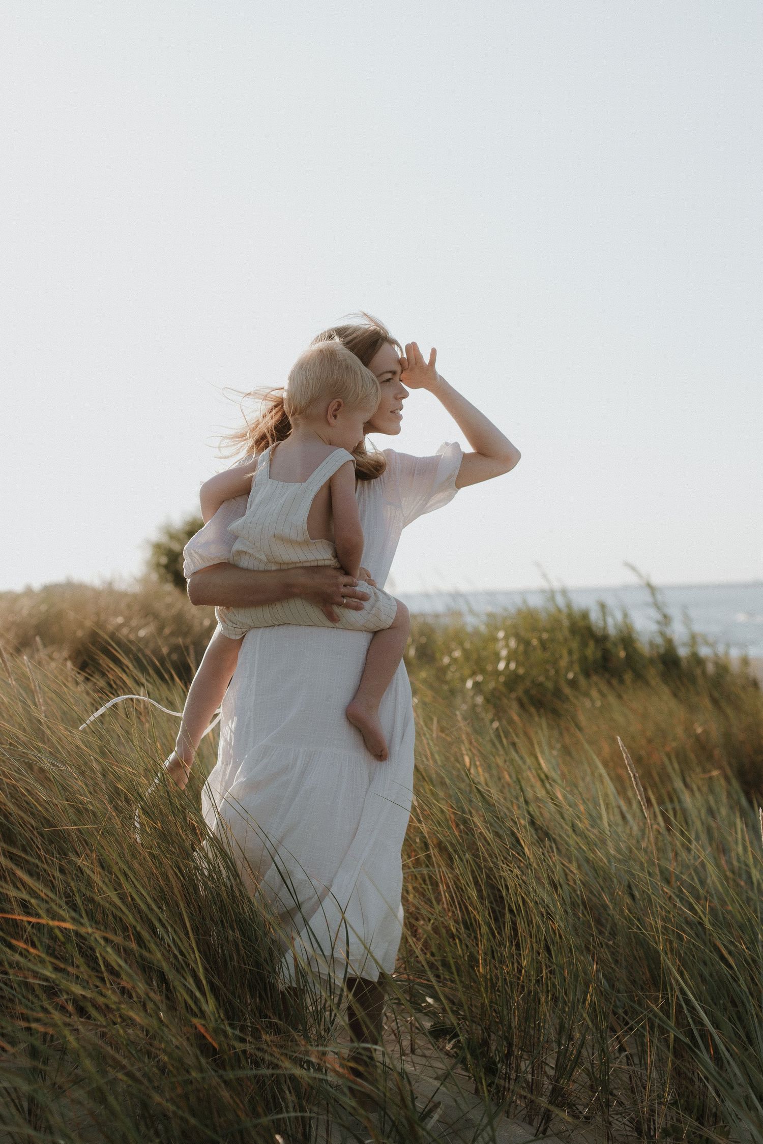 A blonde mother in a white dress carries her toddler through tall beach grass at sunset.