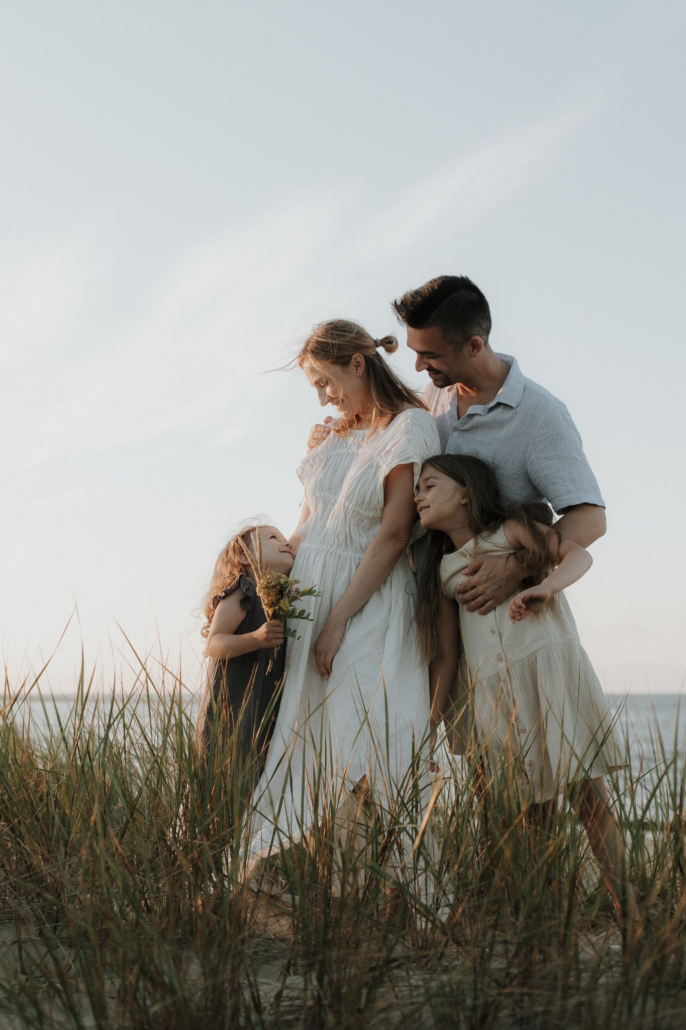 Loving family of four embracing in beach dunes with tall grass at sunset.