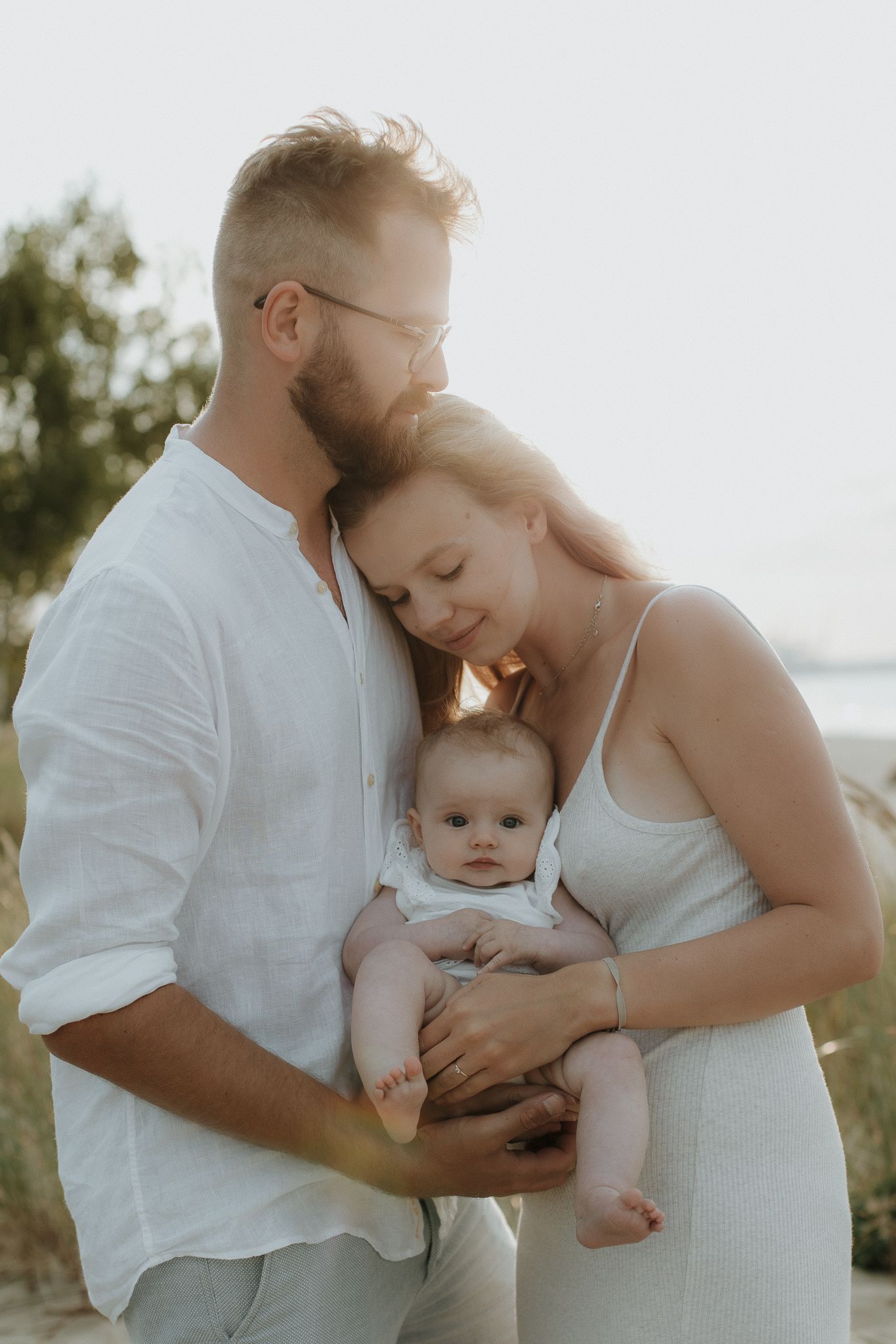 Young couple in white outfits cuddling their infant baby on a sandy beach path at golden hour.