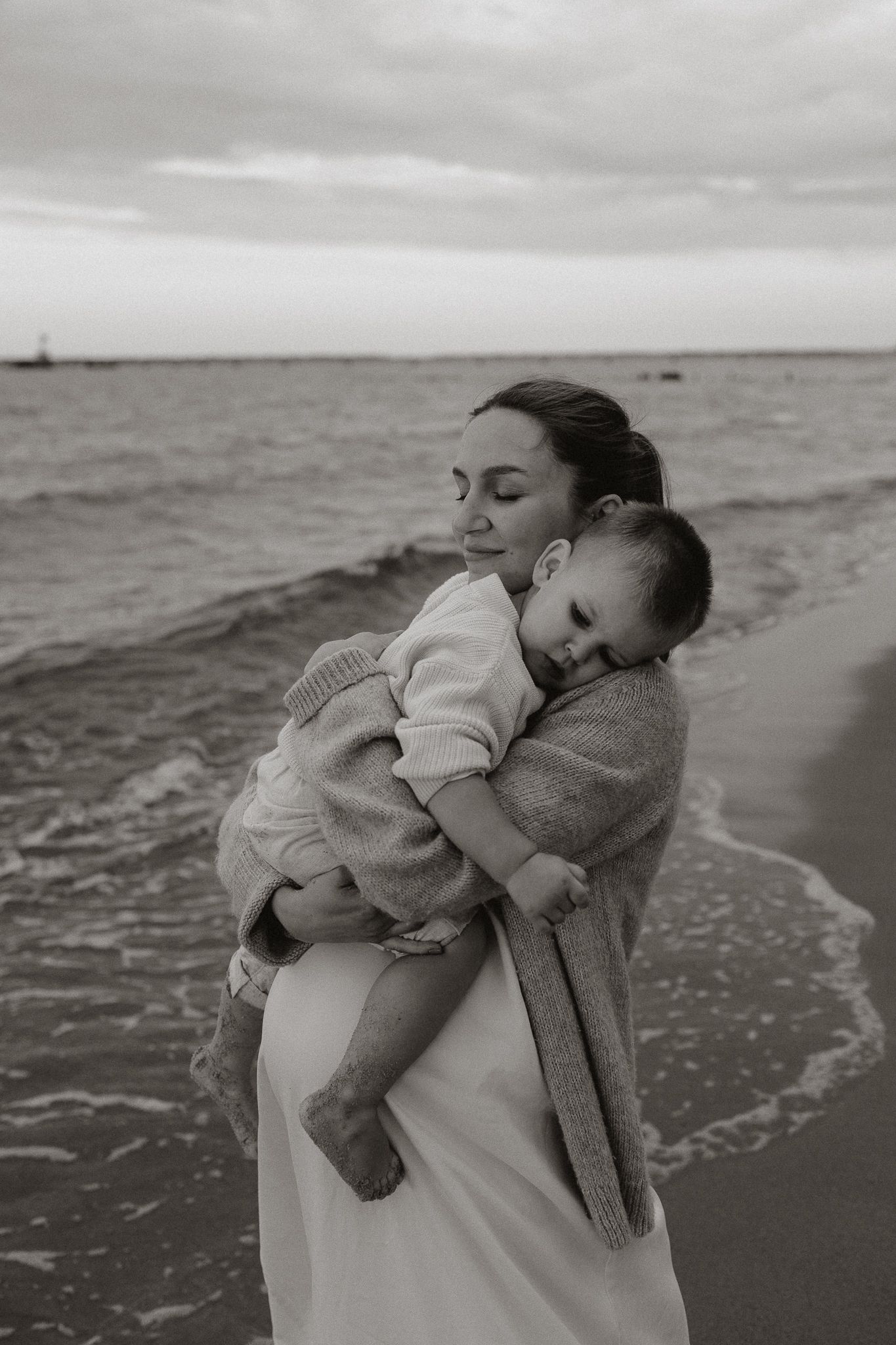 Black and white photo of a mother holding her sleeping baby on a cloudy beach shoreline.
