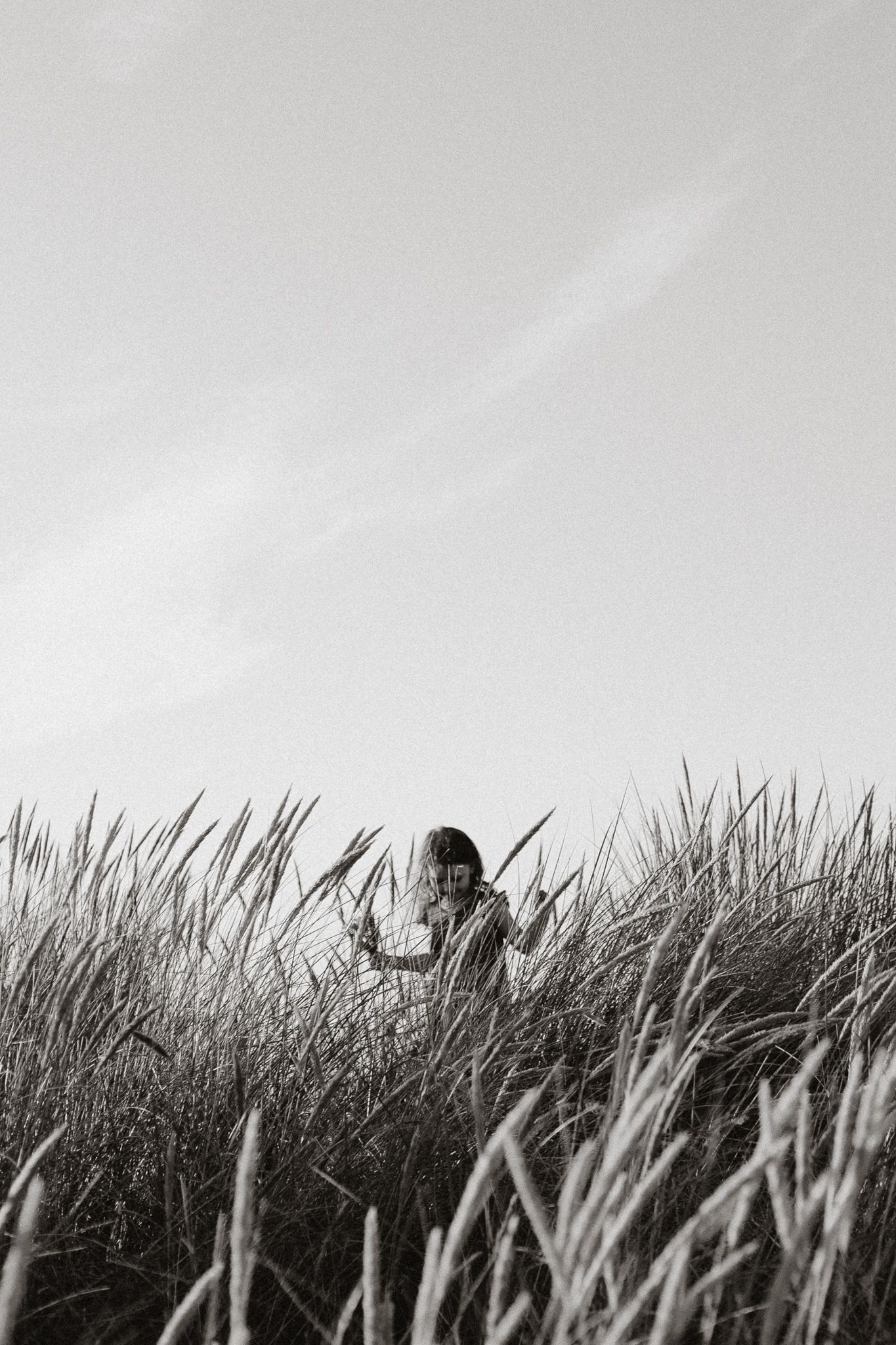 A child peers through tall coastal grass under a wide open sky in black and white.