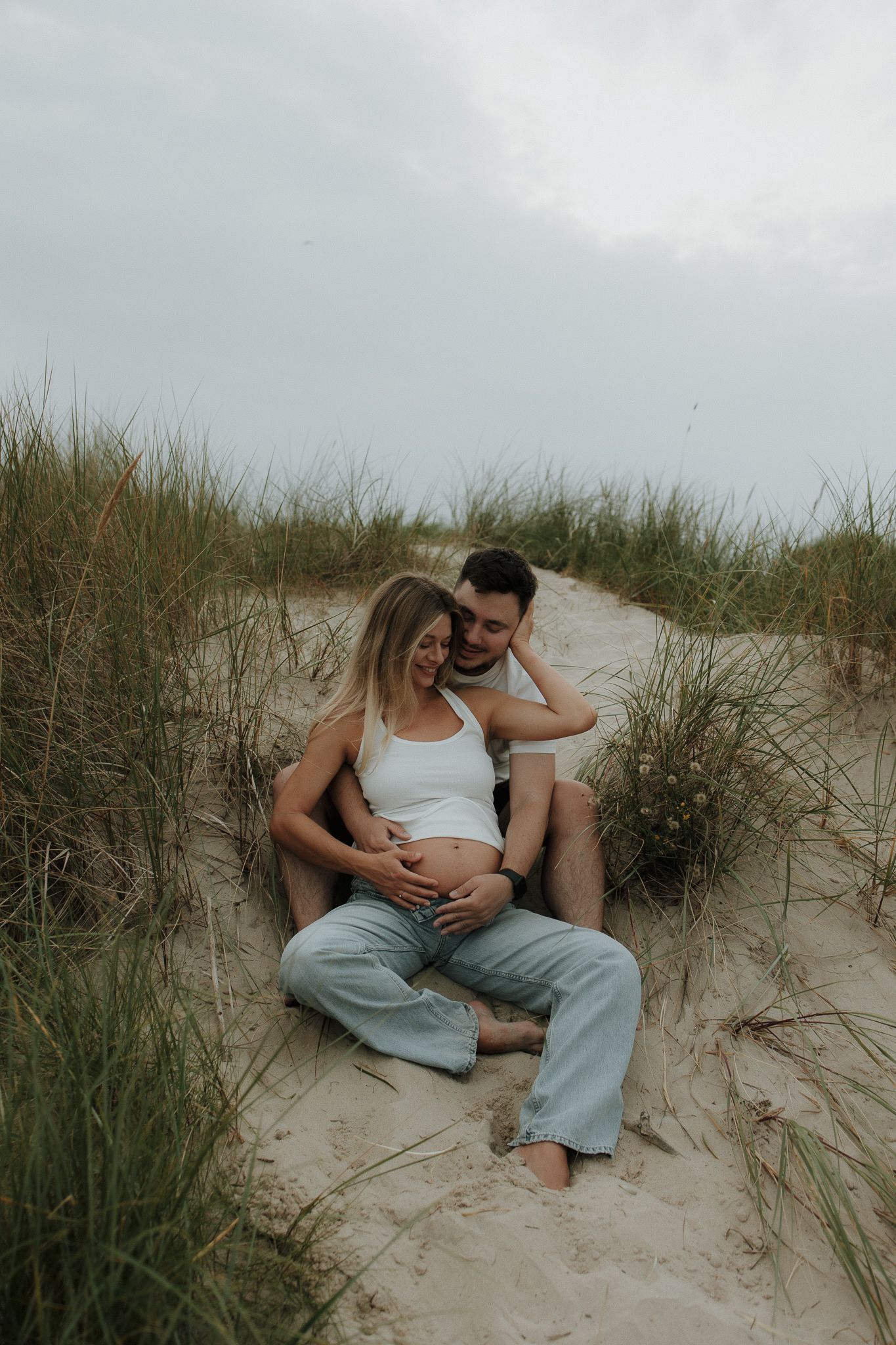 Couple sitting in sand dunes, man embracing pregnant woman from behind on cloudy day.