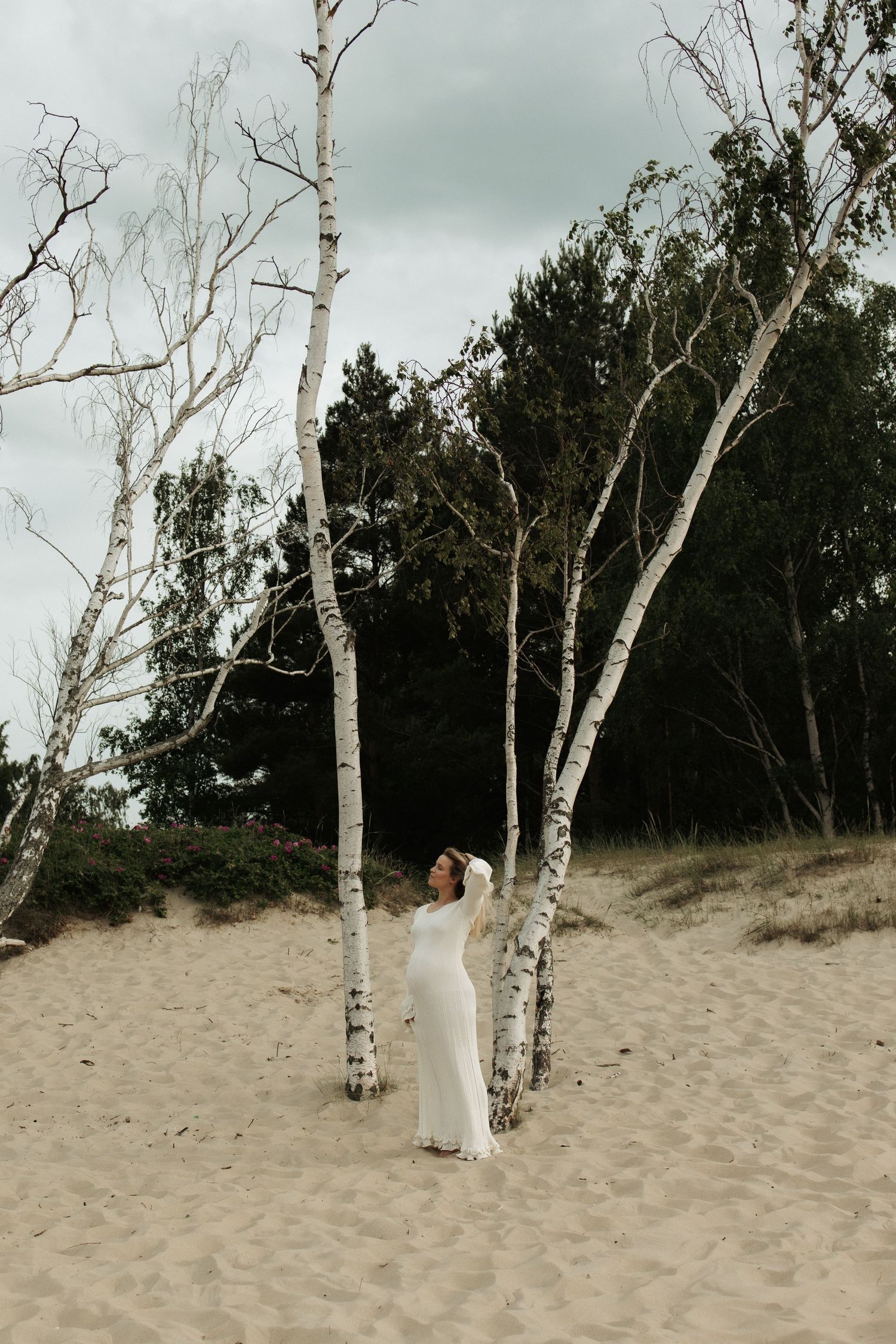 Pregnant woman in white dress stands between birch trees on sandy beach dunes under cloudy sky.