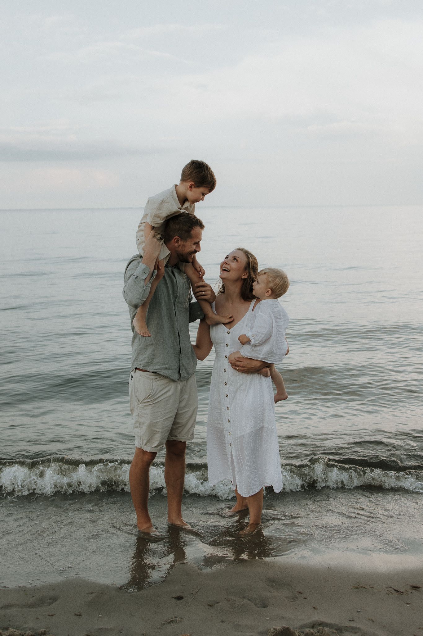 Happy family of four standing in shallow ocean waves at beach during golden hour.