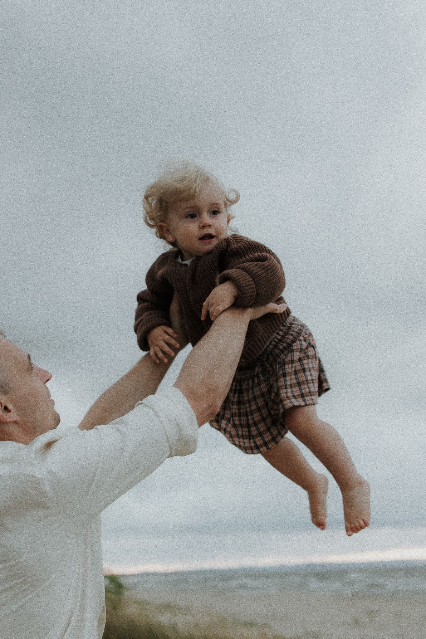 Father lifting happy laughing baby girl high in the air on a cloudy beach day.