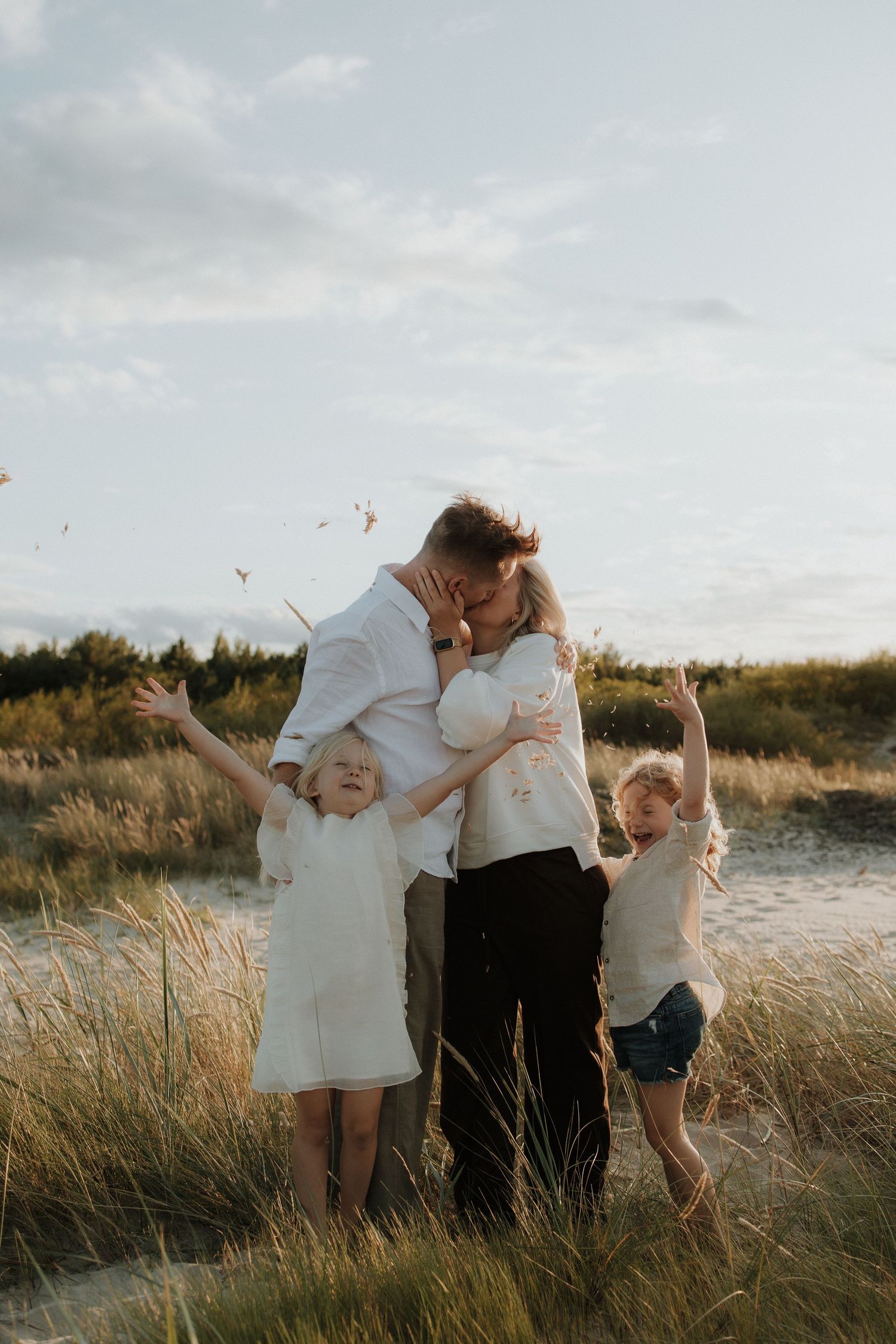 Family of four with parents kissing and children cheering in golden beach grass at sunset.