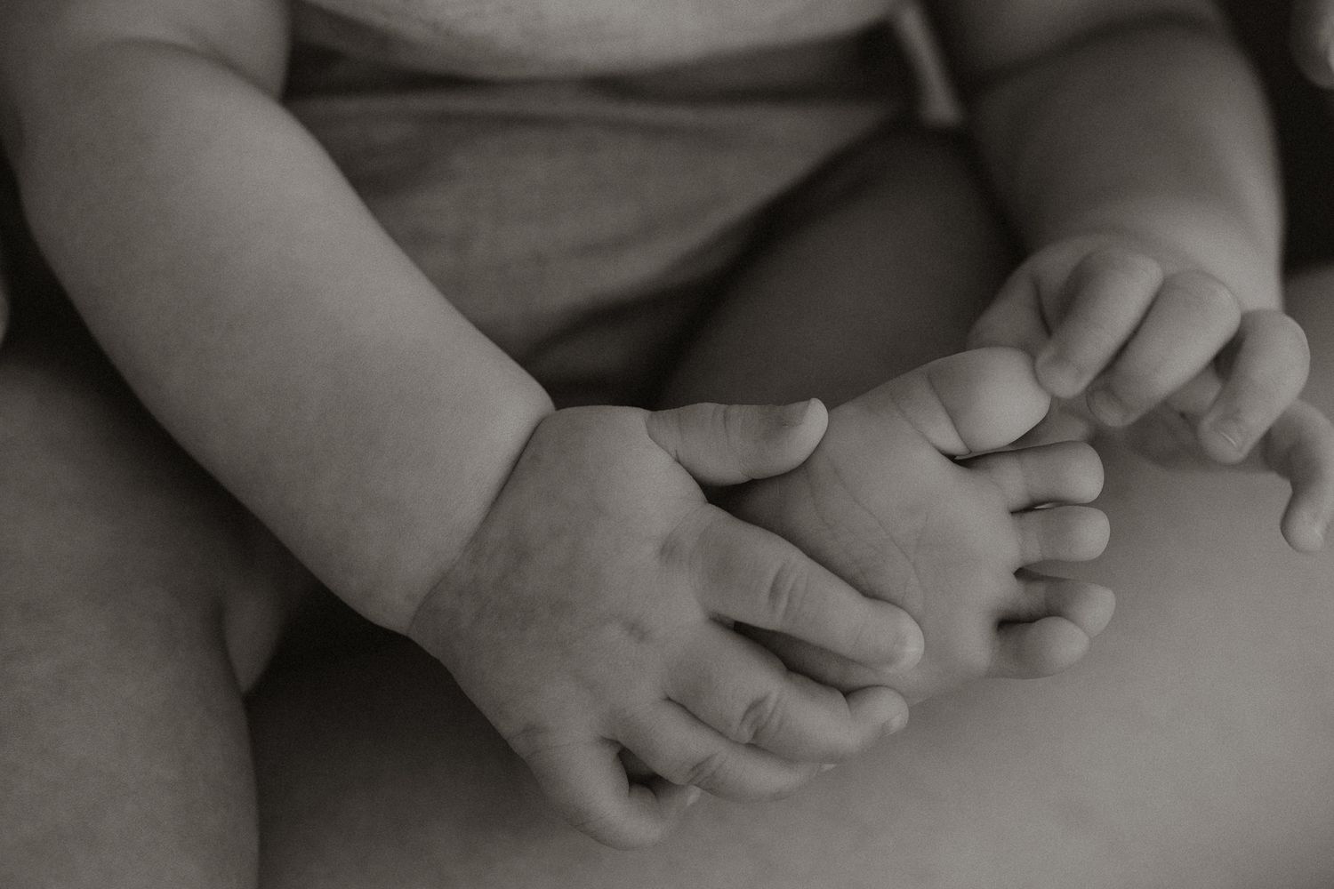 Black and white close-up of a baby grasping their tiny foot with small chubby hands.