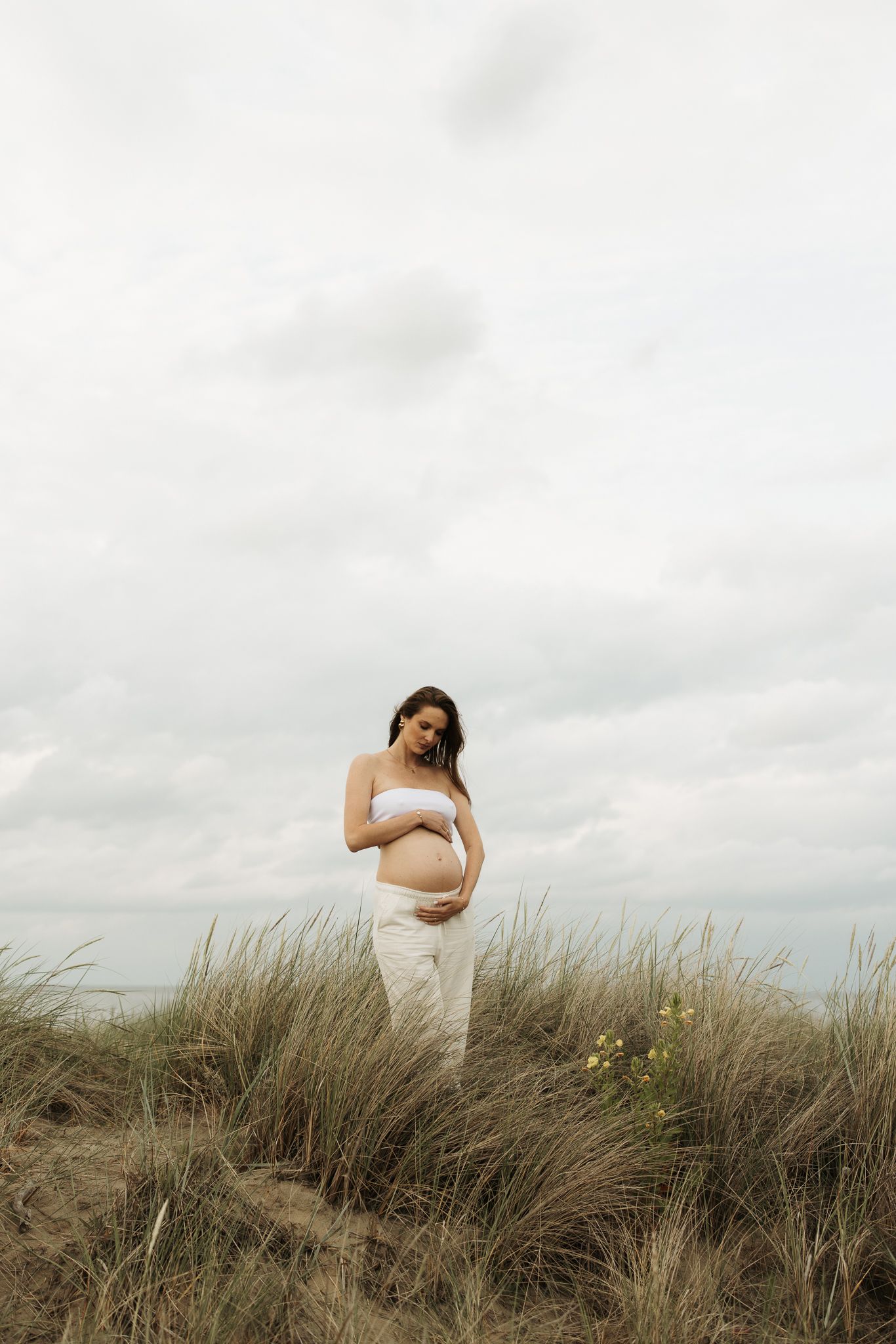 Pregnant woman in white bandeau top holding baby bump in tall coastal grass.