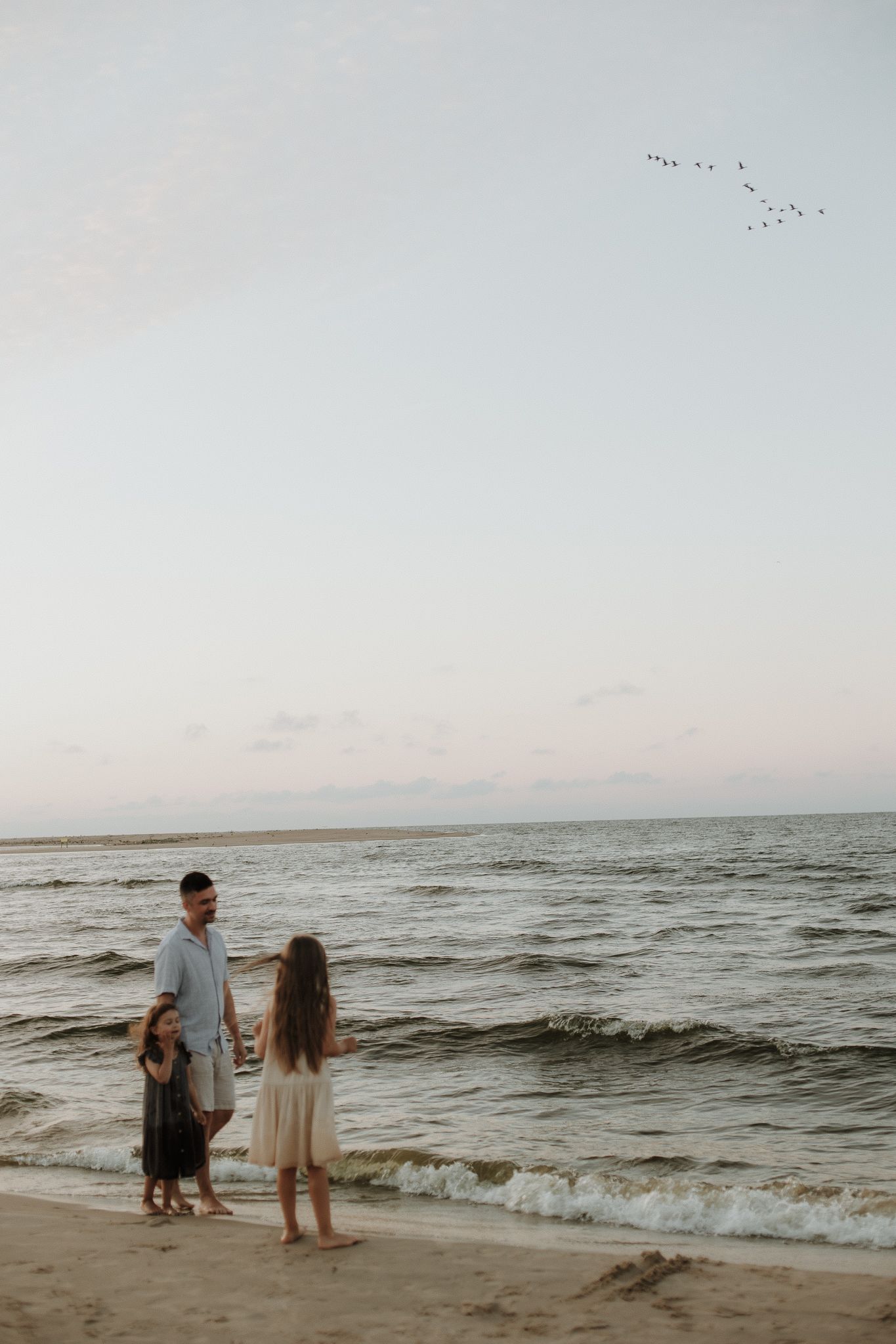 Father standing on a sandy beach at sunset with two young children near ocean waves.