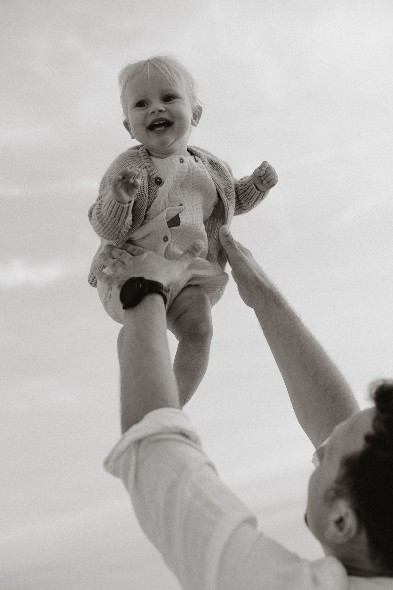 Black and white photo of a father joyfully lifting his laughing baby high into the air outdoors.