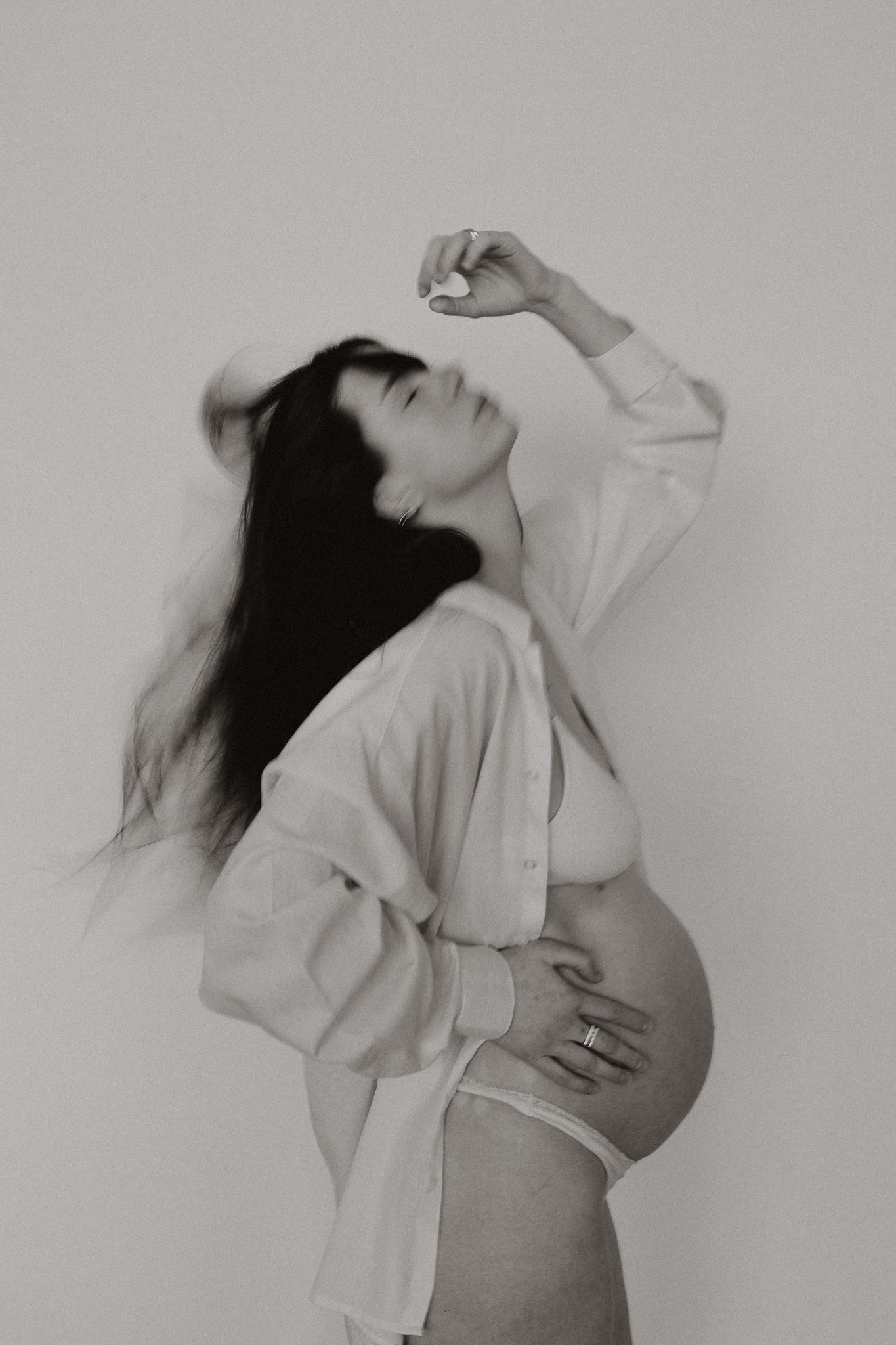Black and white photo of a pregnant woman in white lingerie with flowing hair and one arm raised gracefully.