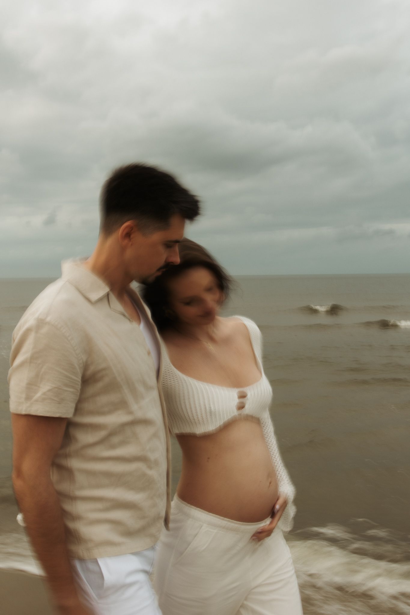 Man and pregnant woman share tender moment on beach with ocean waves behind them.