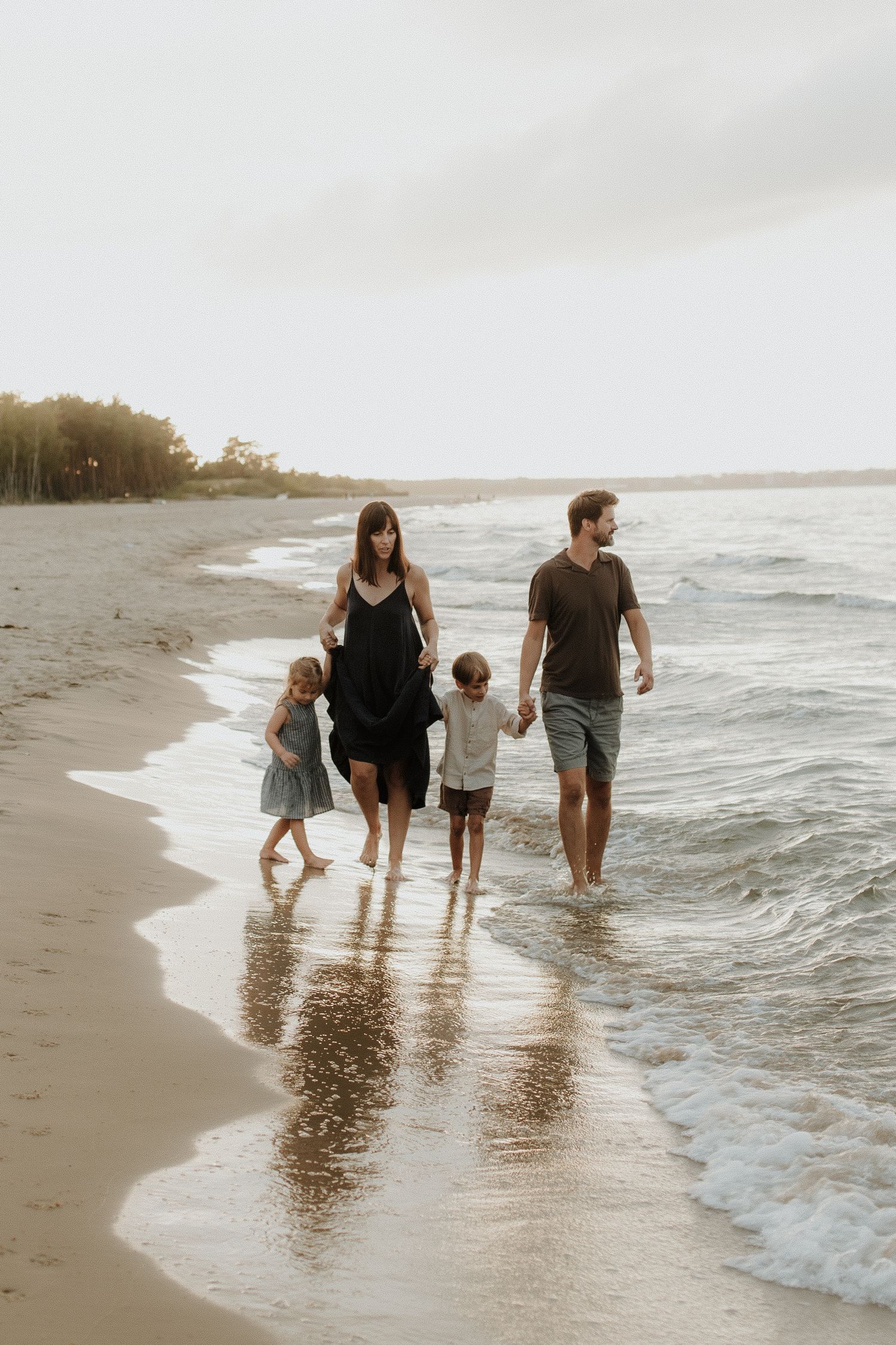 Family of four walking along sandy beach shoreline at sunset, parents holding children's hands near ocean waves.