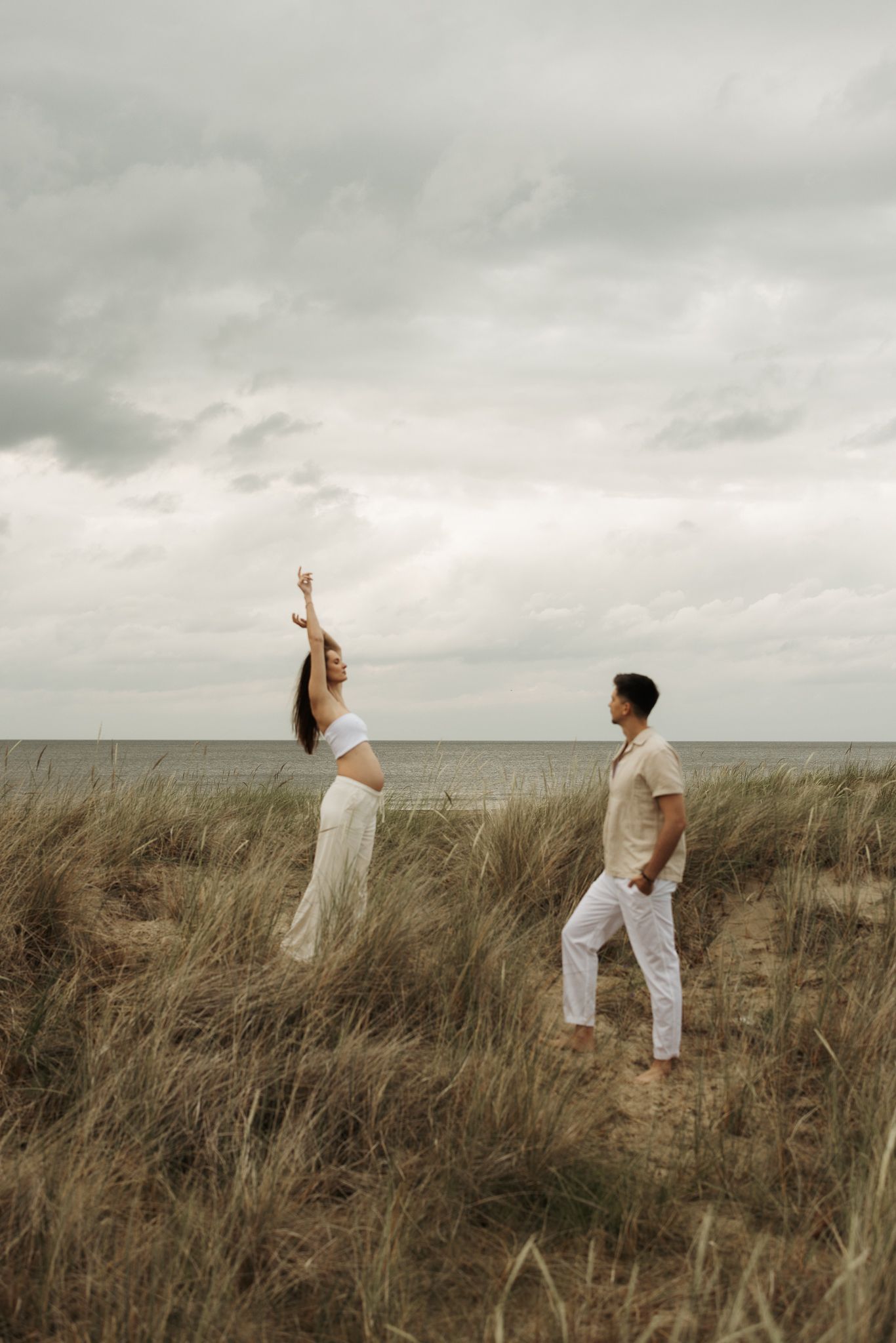 Pregnant woman raising arm joyfully in coastal dunes while partner watches, sea visible in background.