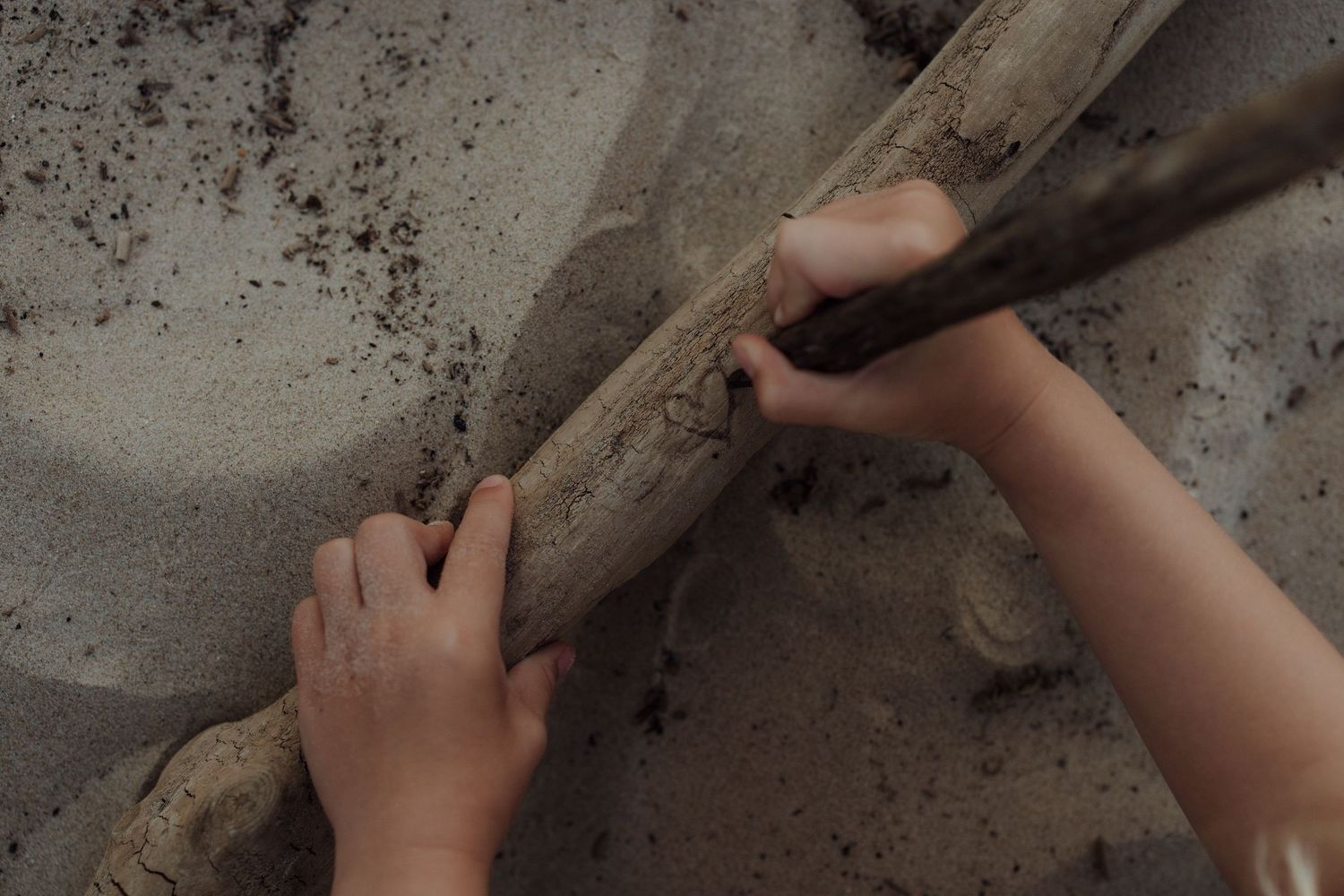 Child's hands playing with a wooden stick in sandy soil outdoors.