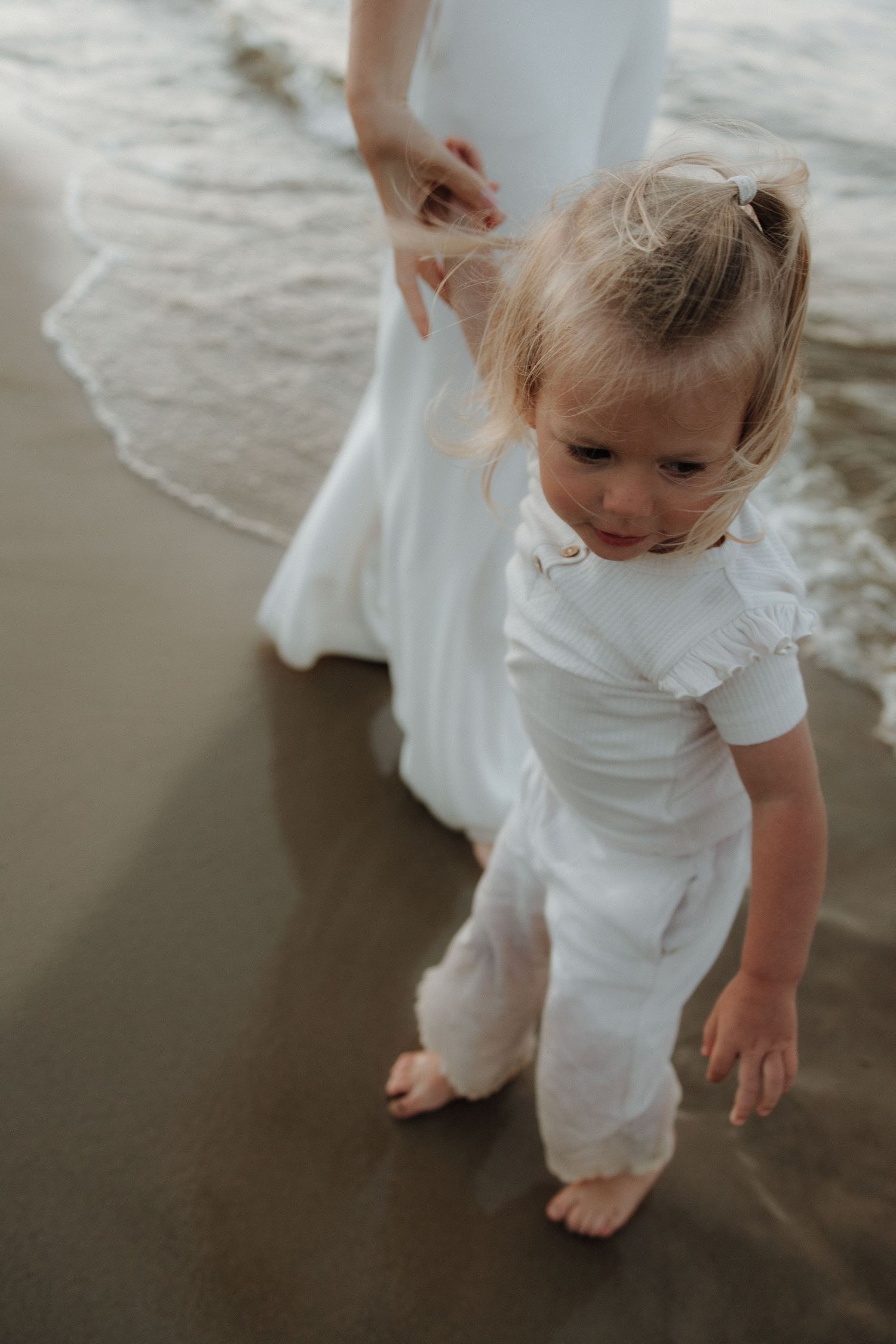 Adorable toddler girl in white outfit walking barefoot on wet sand near ocean waves.