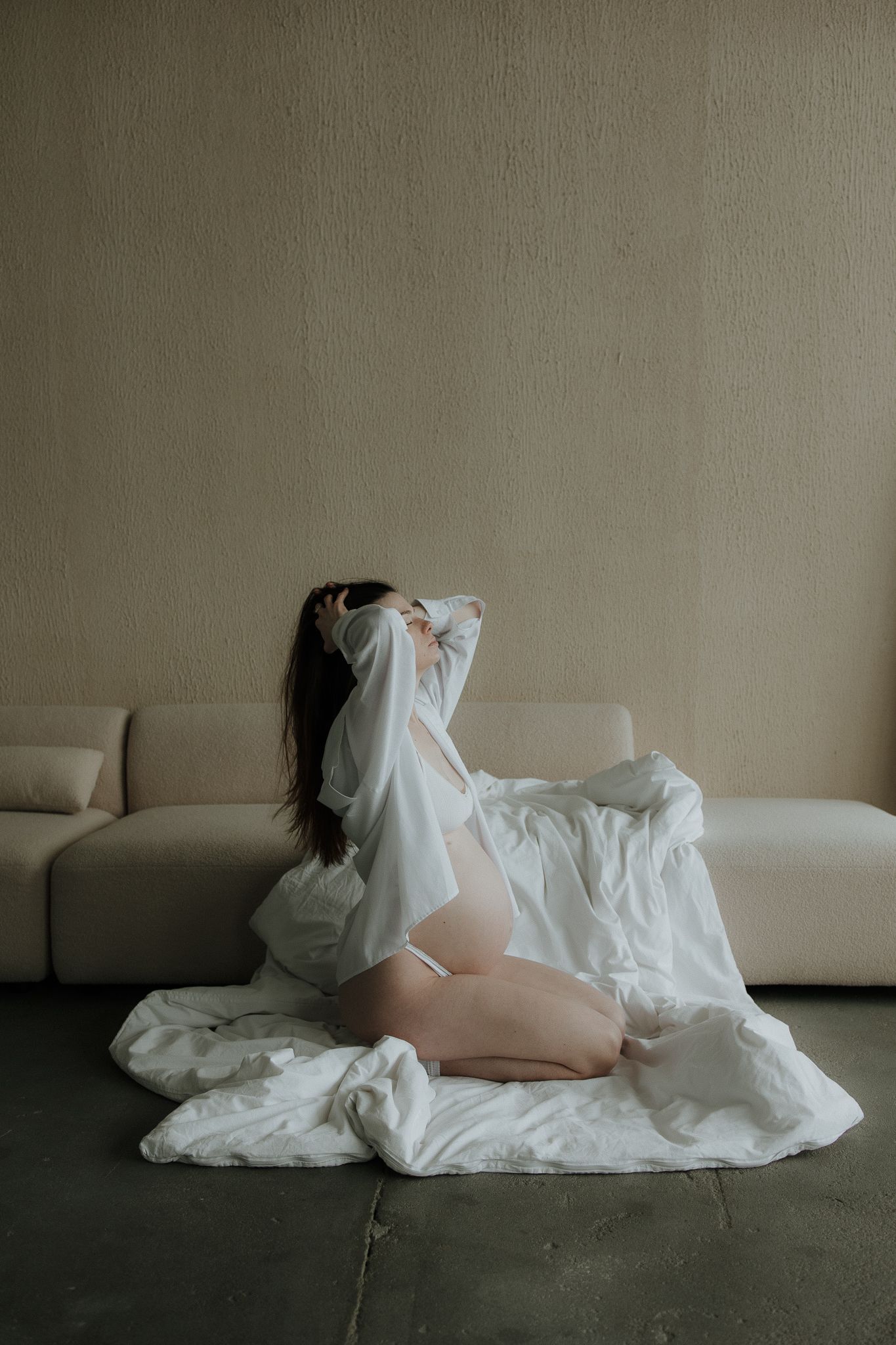 Pregnant woman in white lingerie kneeling beside a beige sofa draped with white sheets, arms raised.