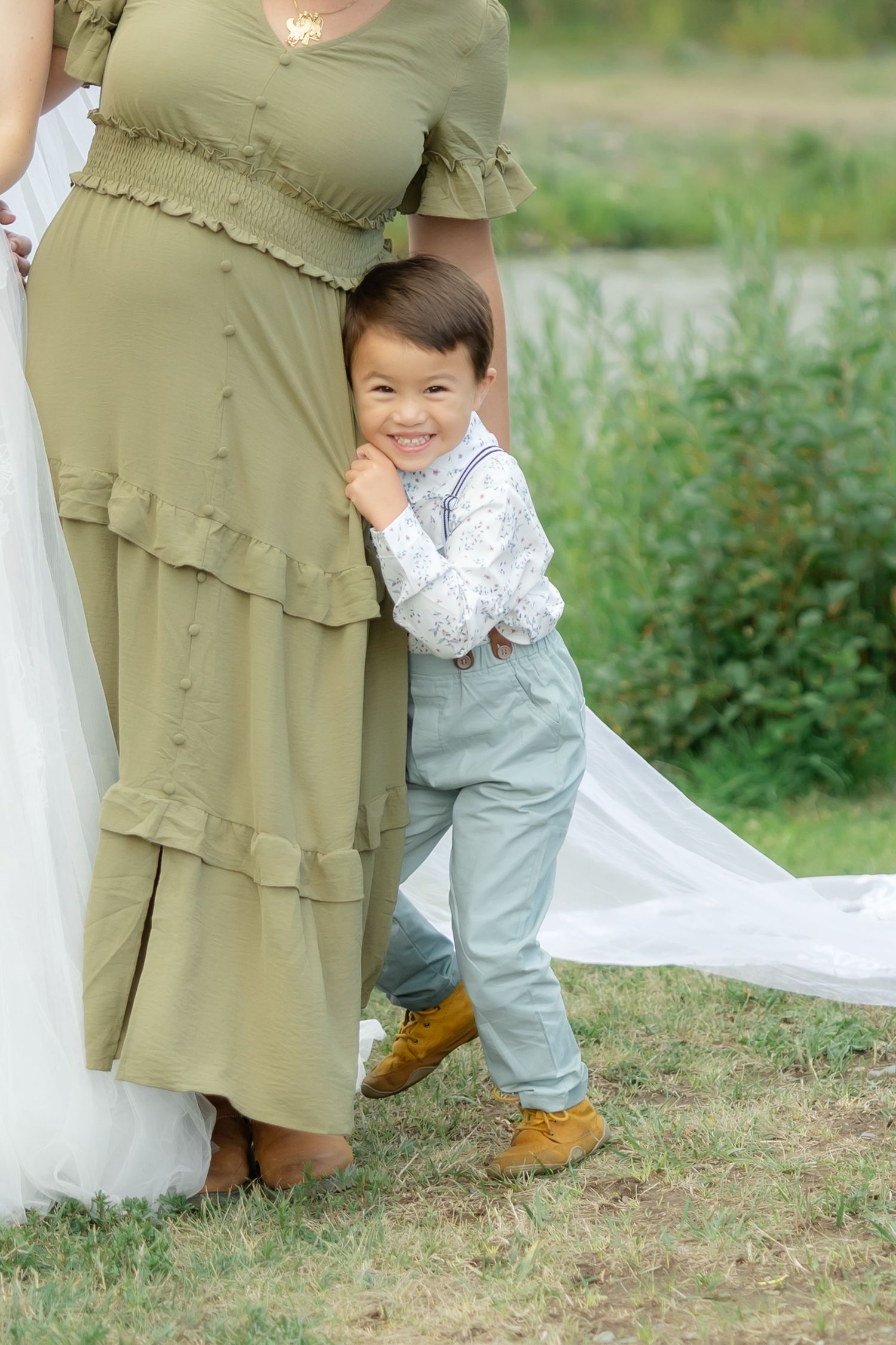 Smiling toddler boy in suspenders hugging a woman in olive dress outdoors at a family photo session.